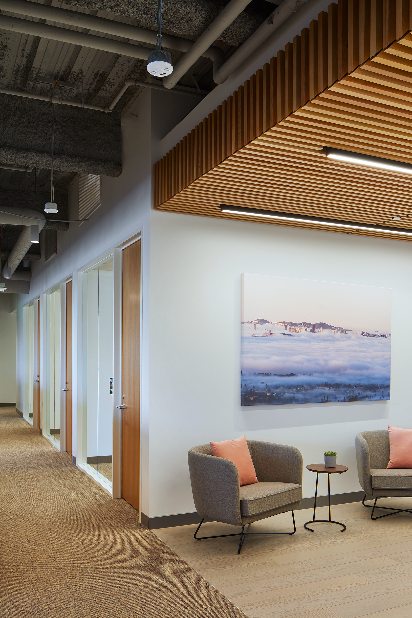Office corridor and lounge area with exposed ceiling systems, wood-slat ceiling detail, glass-fronted rooms, and two upholstered lounge chairs with peach pillows. A large wall photograph hangs above a small side table, creating a quiet seating area beside the hallway.