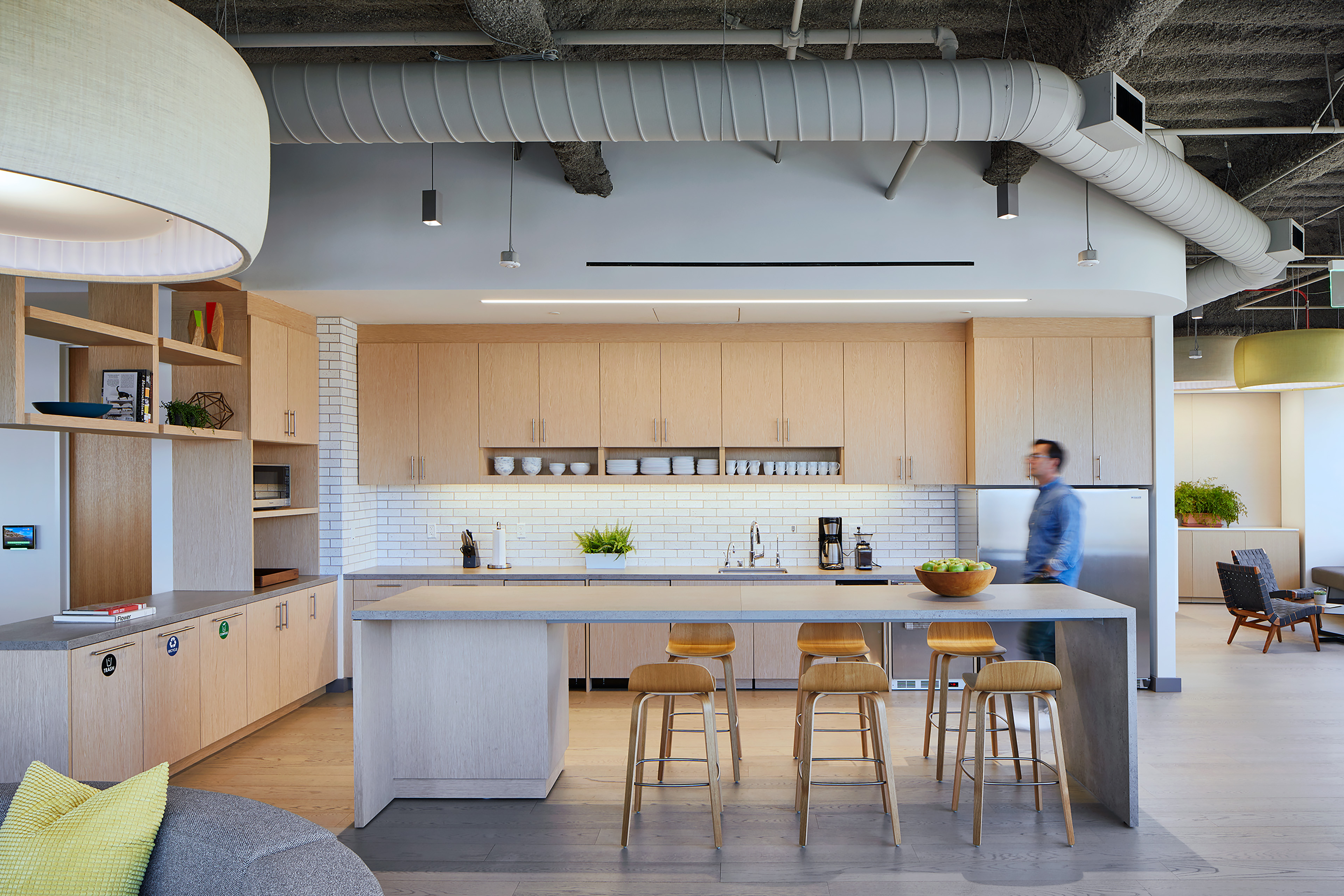Office kitchen and break area with light wood cabinetry, white brick backsplash, stone island, wood stools, and exposed ceiling ducts. A person walks past the counter, with adjacent lounge seating and large pendant lights visible nearby.