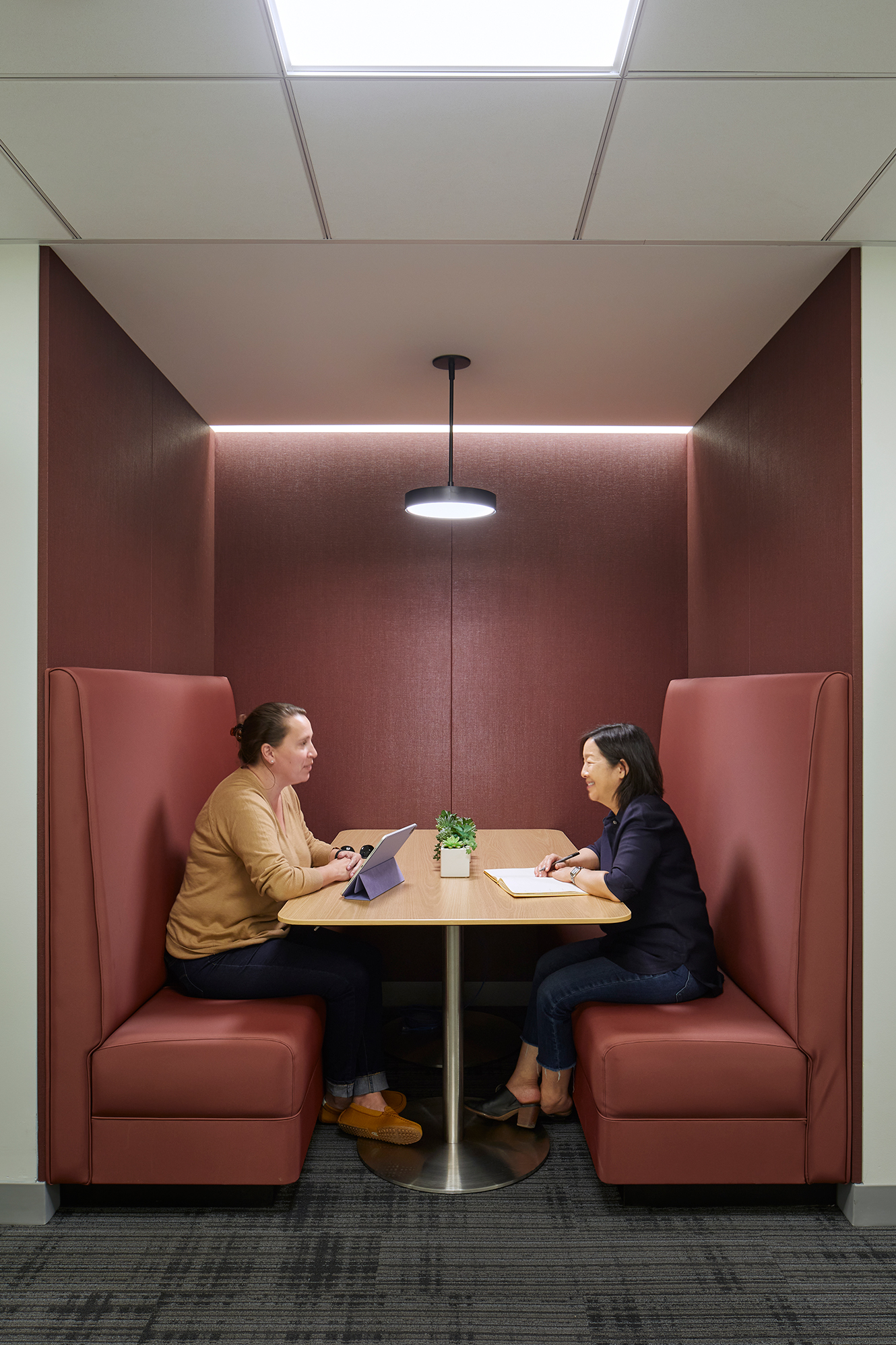 Enclosed meeting booth with red upholstered benches, a small table, and pendant lighting. Two people sit across from each other in conversation, with a tablet, notebook, and small plant on the table.