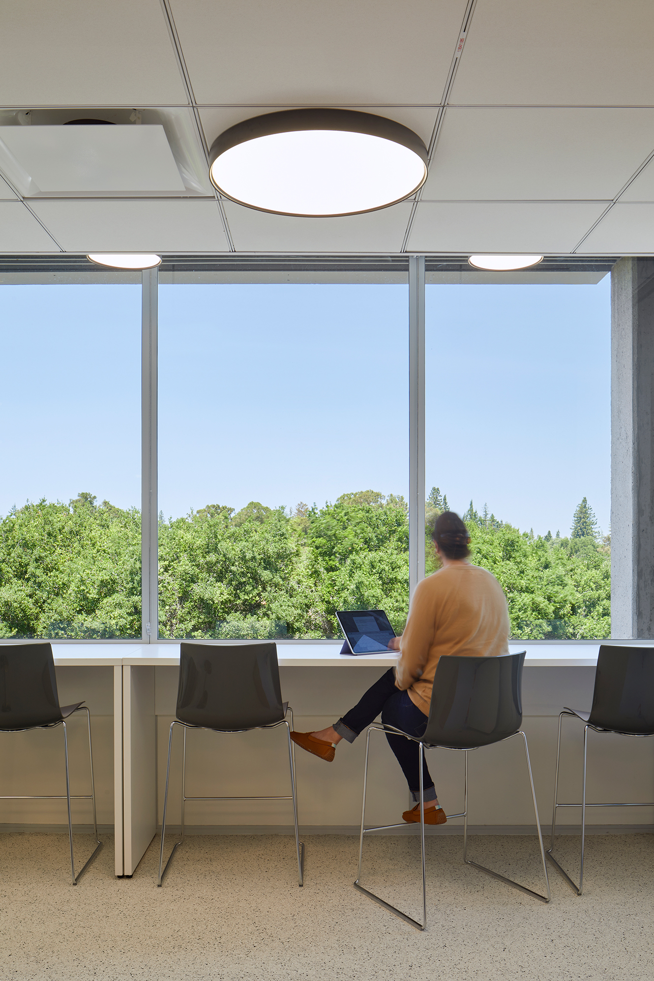 Break room counter seating with large windows overlooking dense trees and a clear sky. A person sits at the window bar with a tablet beneath round pendant lights.