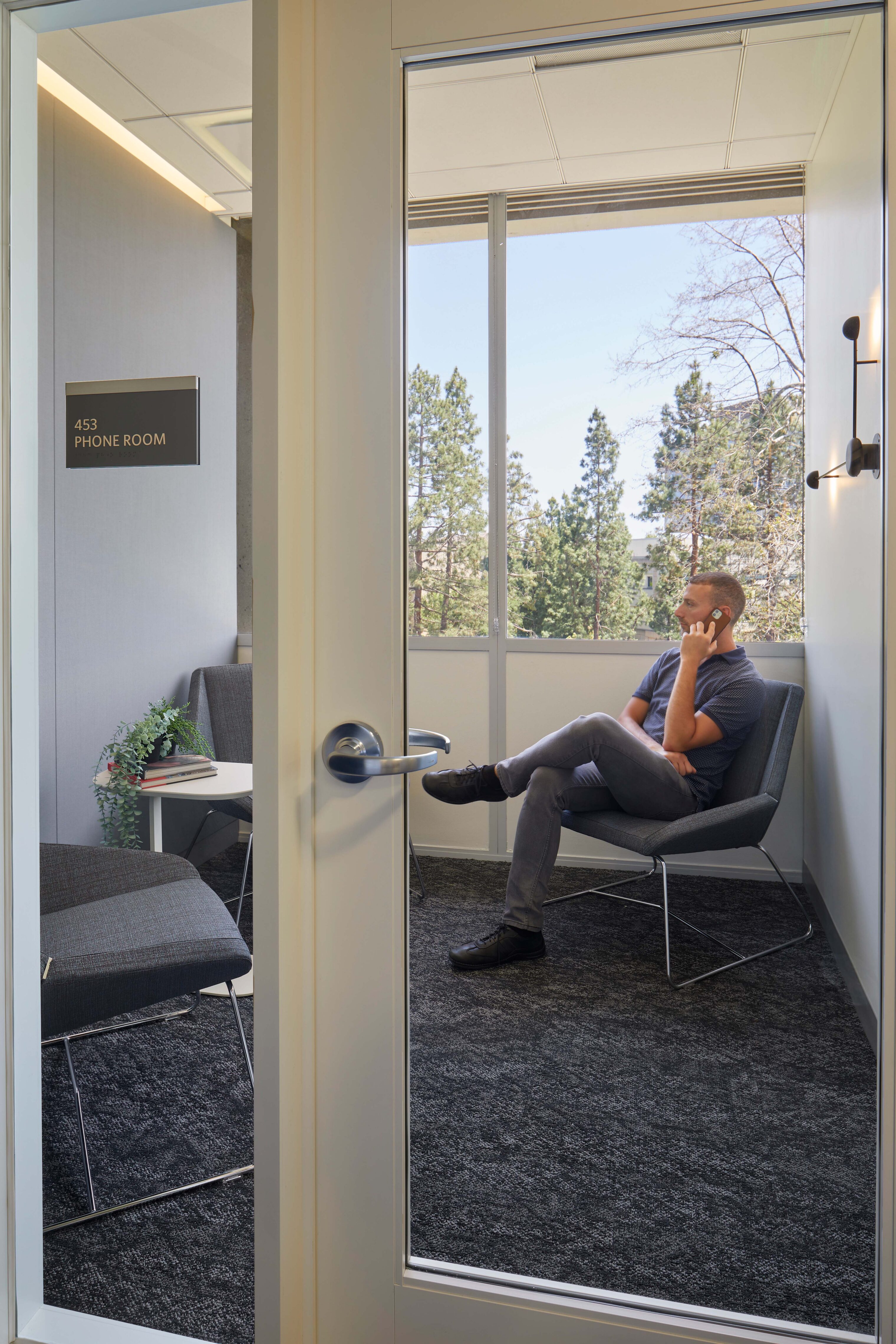 Phone room viewed through a glass door, with a person seated in a lounge chair taking a call. The small private room includes soft seating, a side table with plants and books, wall lighting, and large windows overlooking trees.