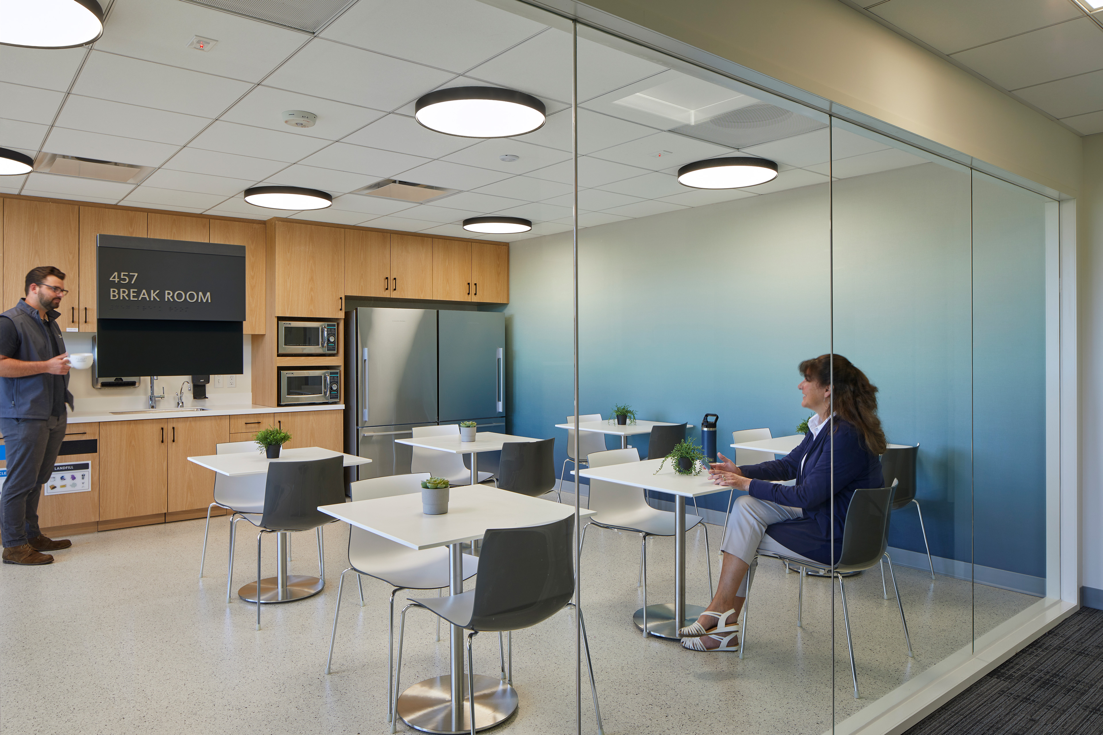 Glass-walled break room with café tables, wood kitchenette cabinetry, microwaves, refrigerators, and a sink. Two people use the shared space, which includes small plants, round pendant lights, and a sign reading “457 Break Room.”