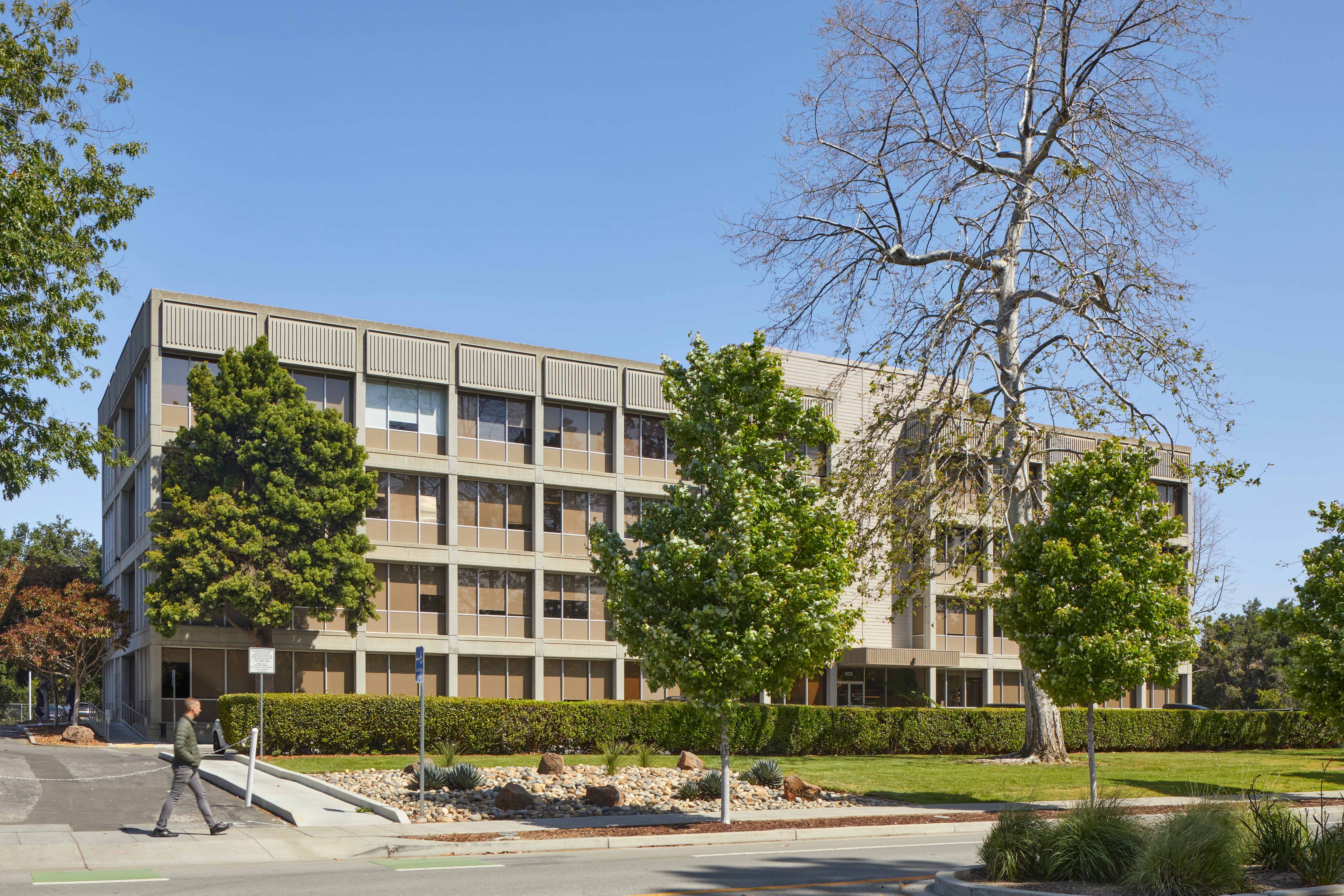xterior view of a multi-story office building set behind landscaped grounds, trees, and a sidewalk. A pedestrian walks past the building on a sunny day under a clear blue sky.