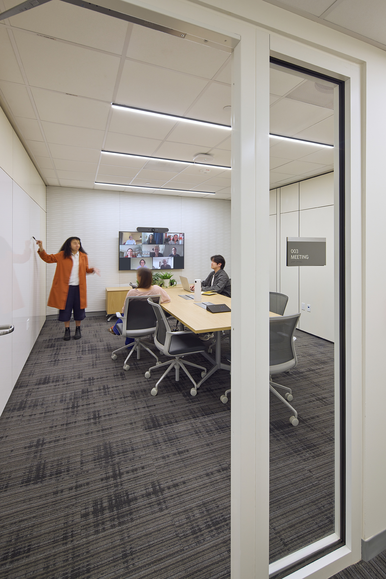 Bright open office with low workstations, built-in storage, and floor-to-ceiling windows overlooking trees. Two employees converse near the windows while others work nearby, with glass-fronted meeting rooms lining the opposite side.