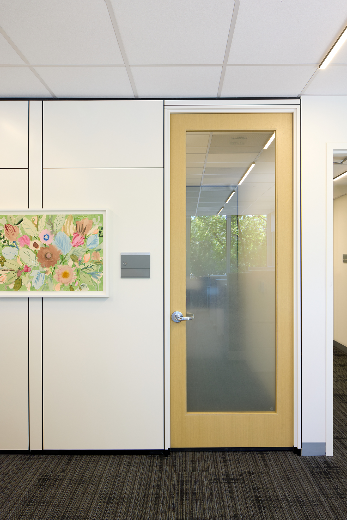 Office corridor wall with a wood-framed glass door labeled “216” and a colorful floral artwork mounted beside it. The door’s frosted lower glass provides privacy while allowing light and views from the office beyond.