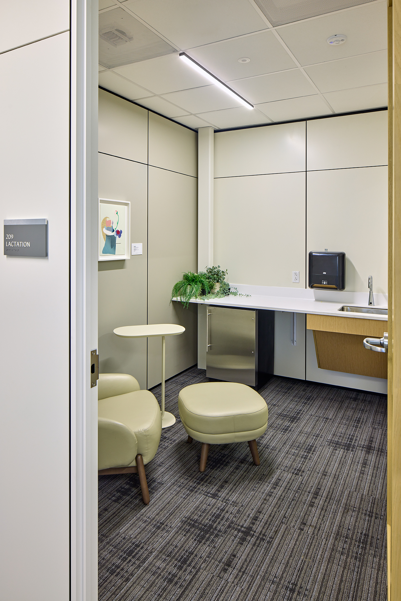 Lactation room with a lounge chair, ottoman, small side table, sink, undercounter refrigerator, plants, and wall-mounted artwork. The private room is viewed through an open door with a sign reading “209 Lactation.”