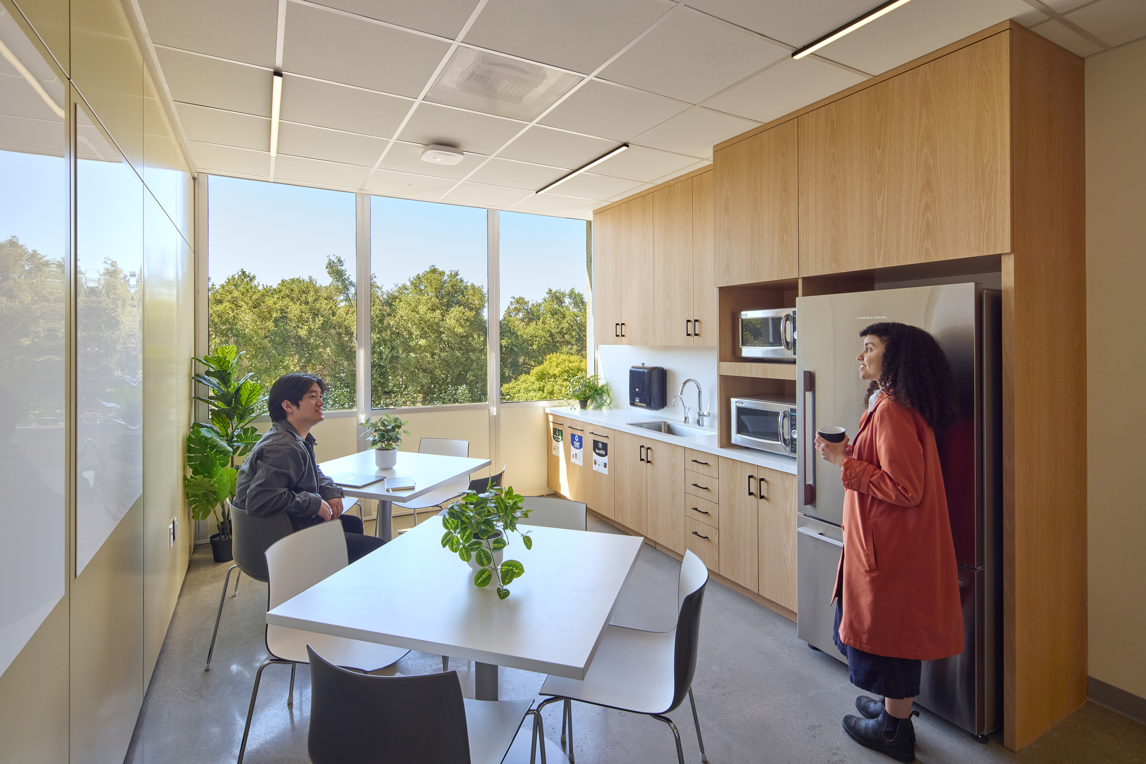 Break room with wood cabinetry, tables, plants, and large windows overlooking trees. Two employees talk near the kitchenette, which includes a sink, microwaves, refrigerator, and waste sorting bins.