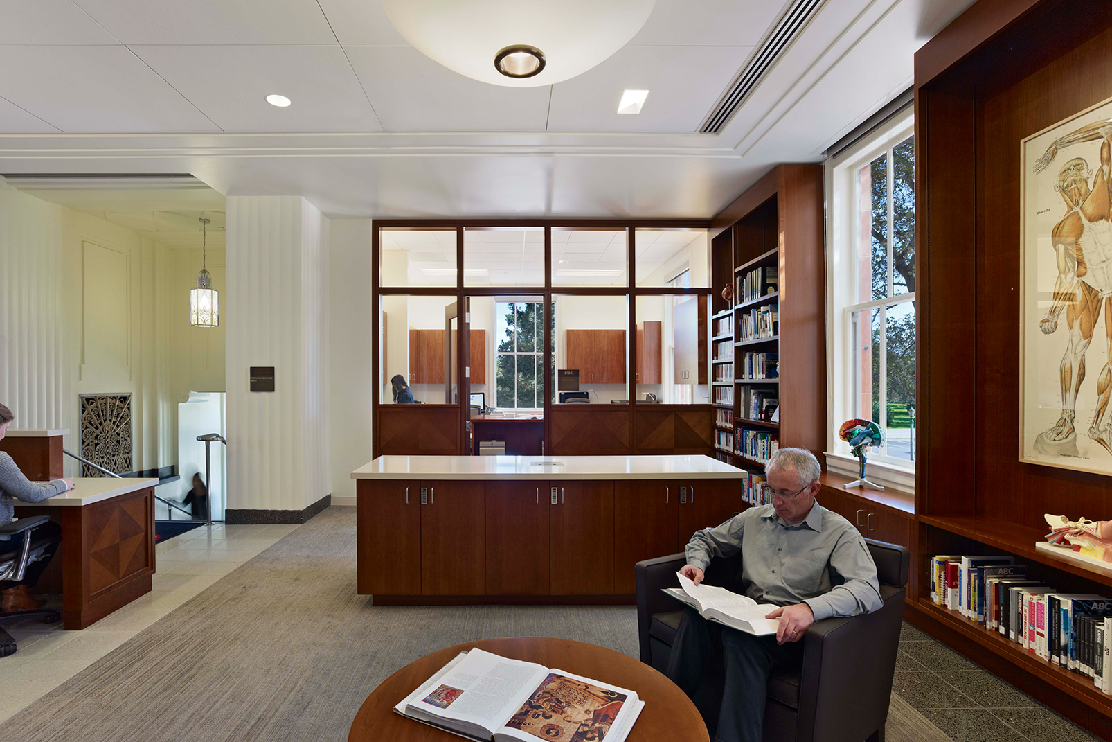Healthcare resource and consultation area with wood cabinetry, built-in bookshelves, anatomical displays, and people reading or working nearby.
