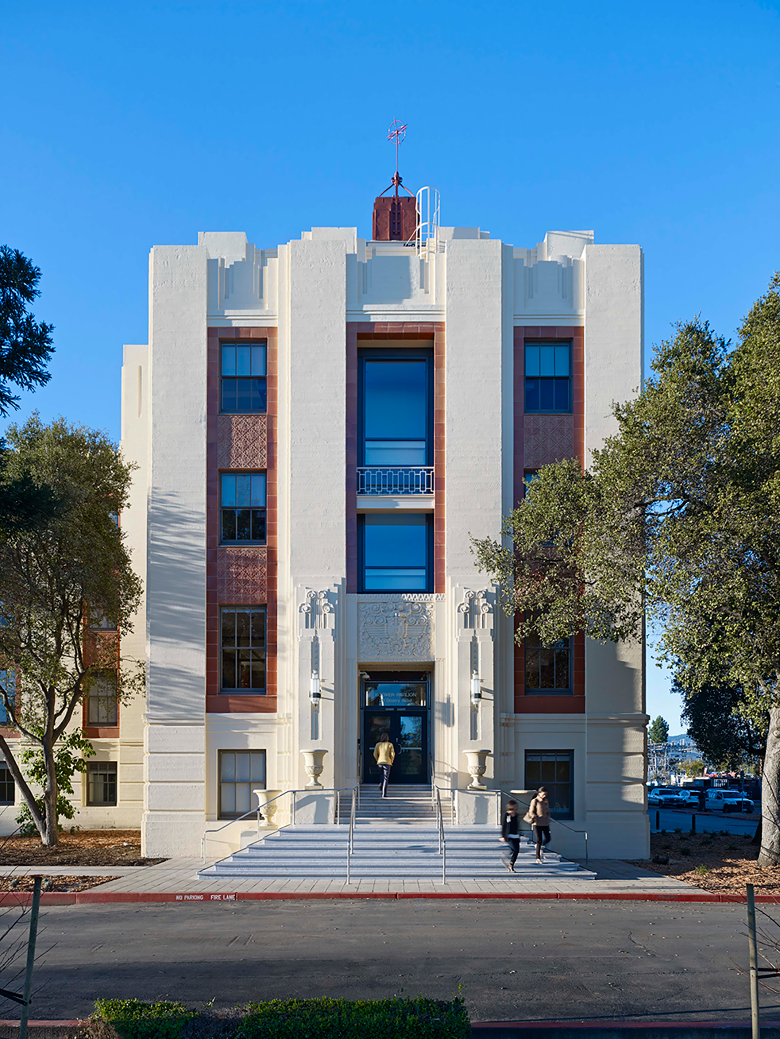 Front entrance of a restored Art Deco building with broad steps, decorative masonry, and people entering and leaving beneath a clear blue sky.