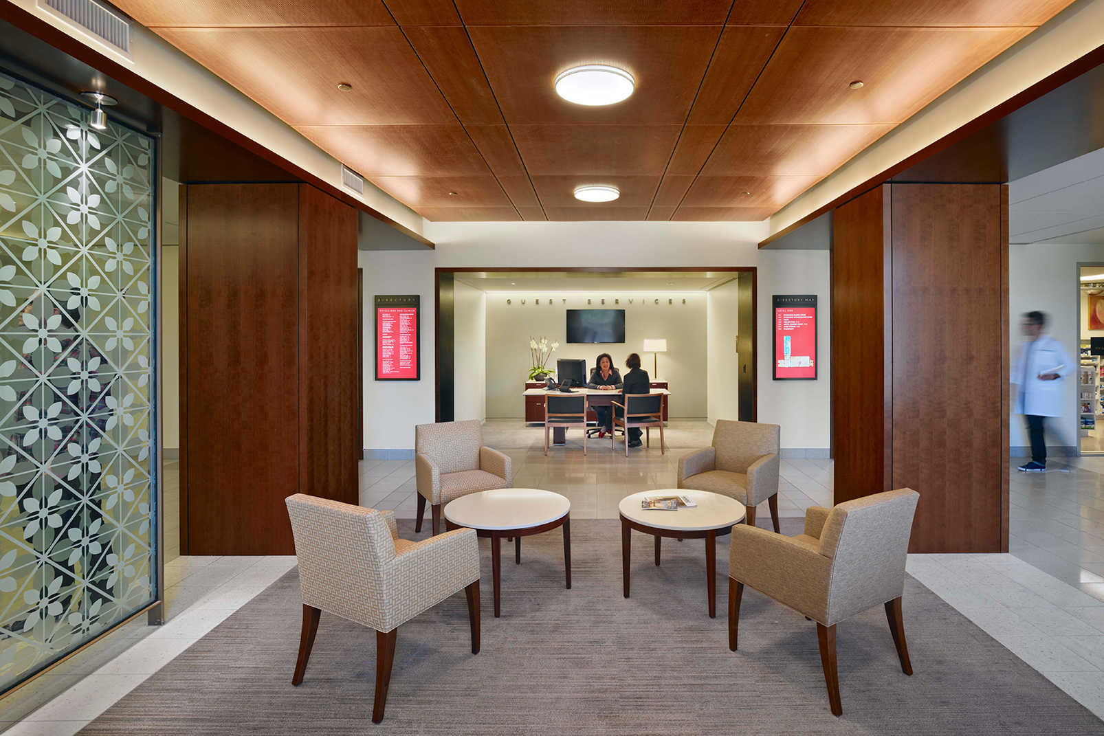 Healthcare lobby with warm wood paneling, patterned glass screens, upholstered seating, and a guest services desk in the background.