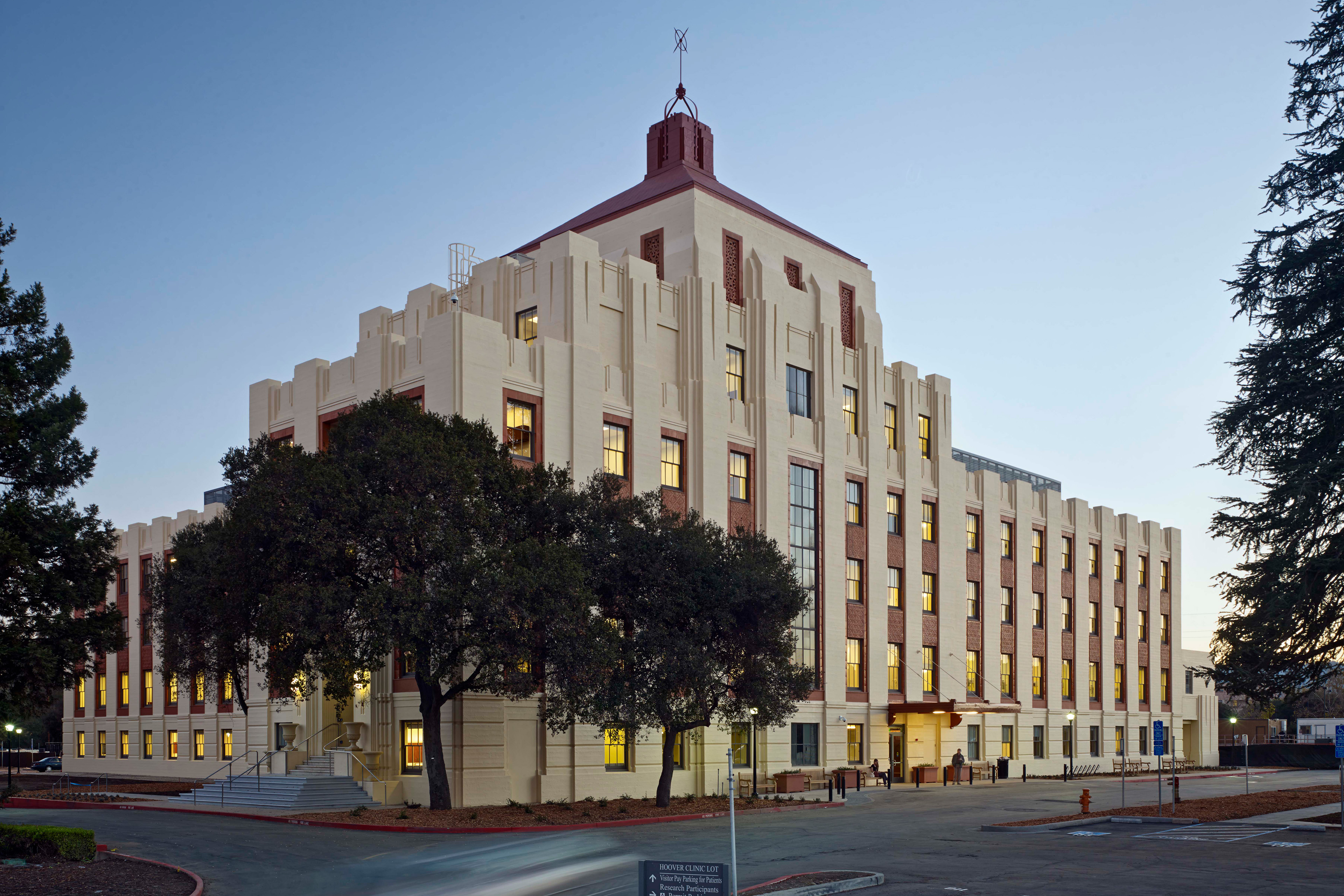 Historic multi-story Art Deco building at dusk, with cream and red masonry, illuminated windows, mature trees, and a clear blue sky.