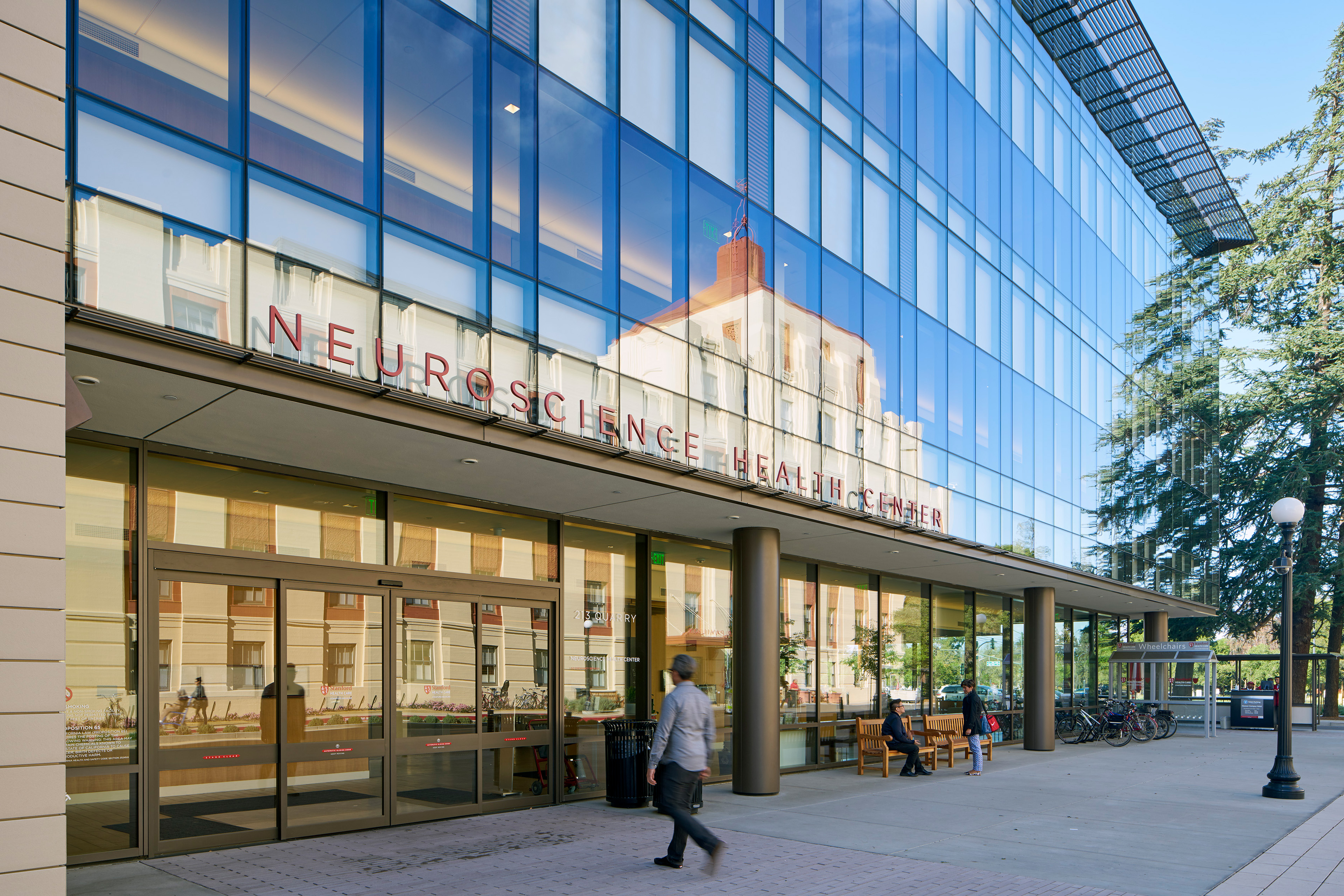 Main entrance to the Neuroscience Health Center with a reflective glass façade, entrance canopy, benches, bicycle parking, and visitors moving through the plaza.