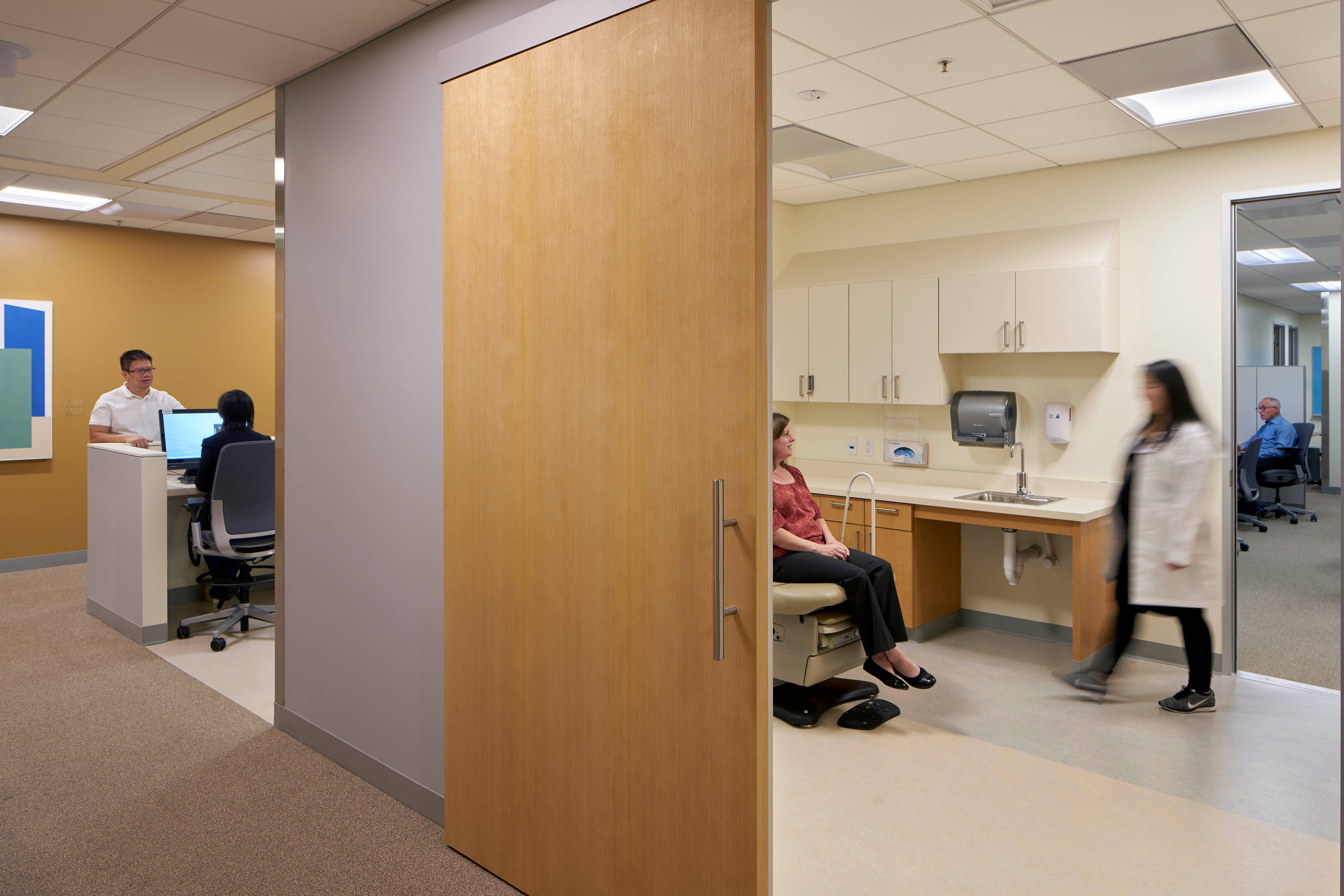Clinical exam area with a large sliding wood door, a patient seated beside an accessible sink, a clinician walking through the room, and staff working at adjacent open workstations.