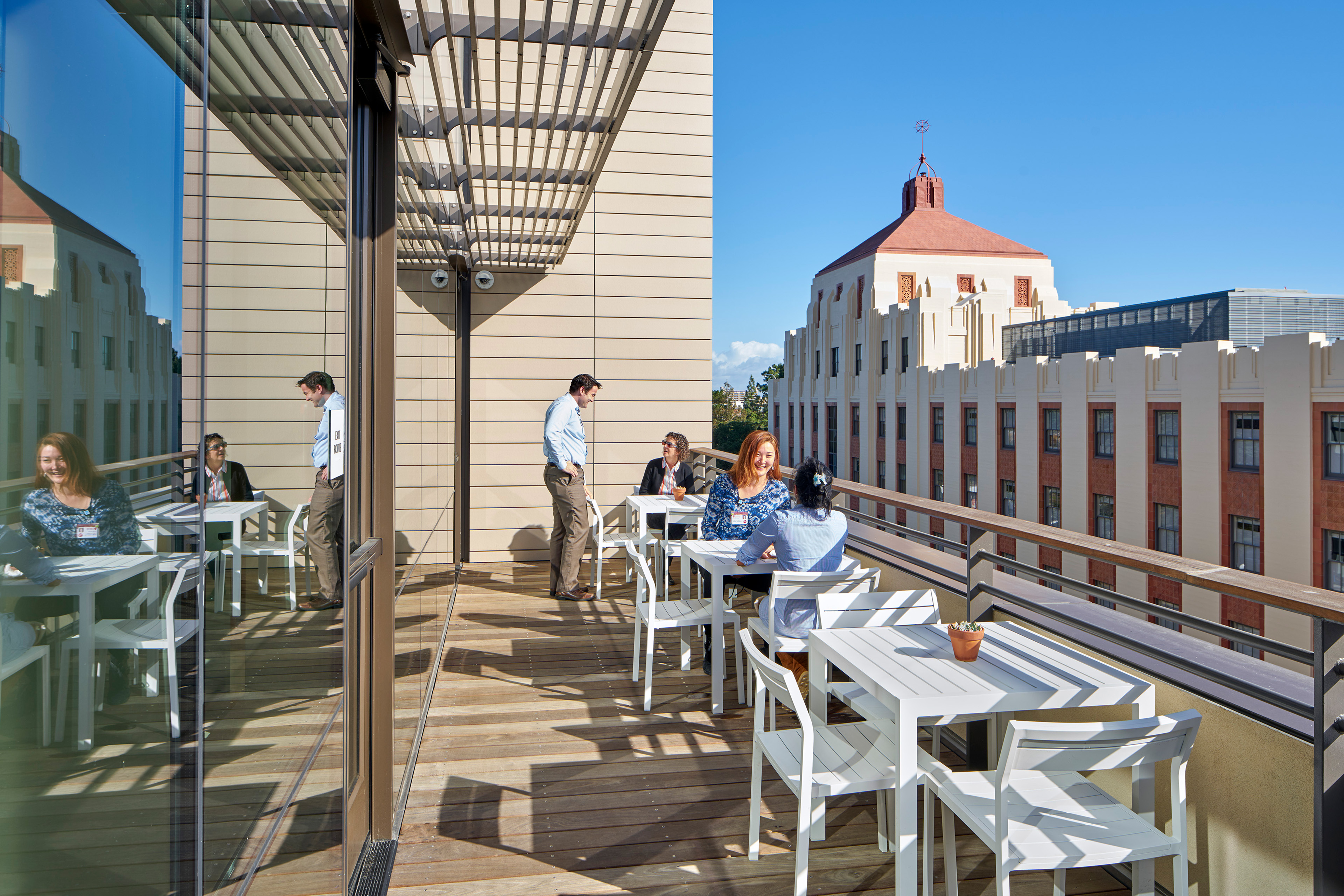 Outdoor terrace at the Neuroscience Health Center with white tables and chairs, staff and visitors gathering, and views of the adjacent historic hospital building.