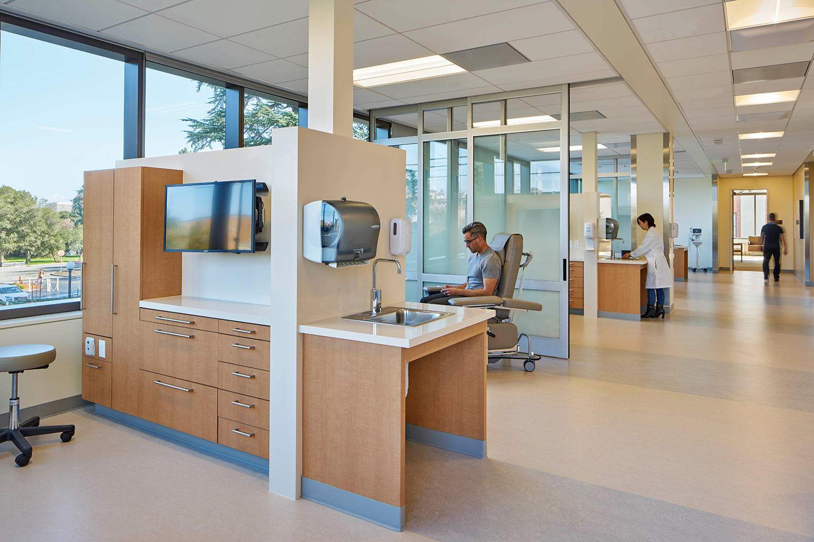 Bright outpatient treatment area with wood cabinetry, an accessible sink, glass-enclosed care bays, large windows, and patients and clinical staff using the space.
