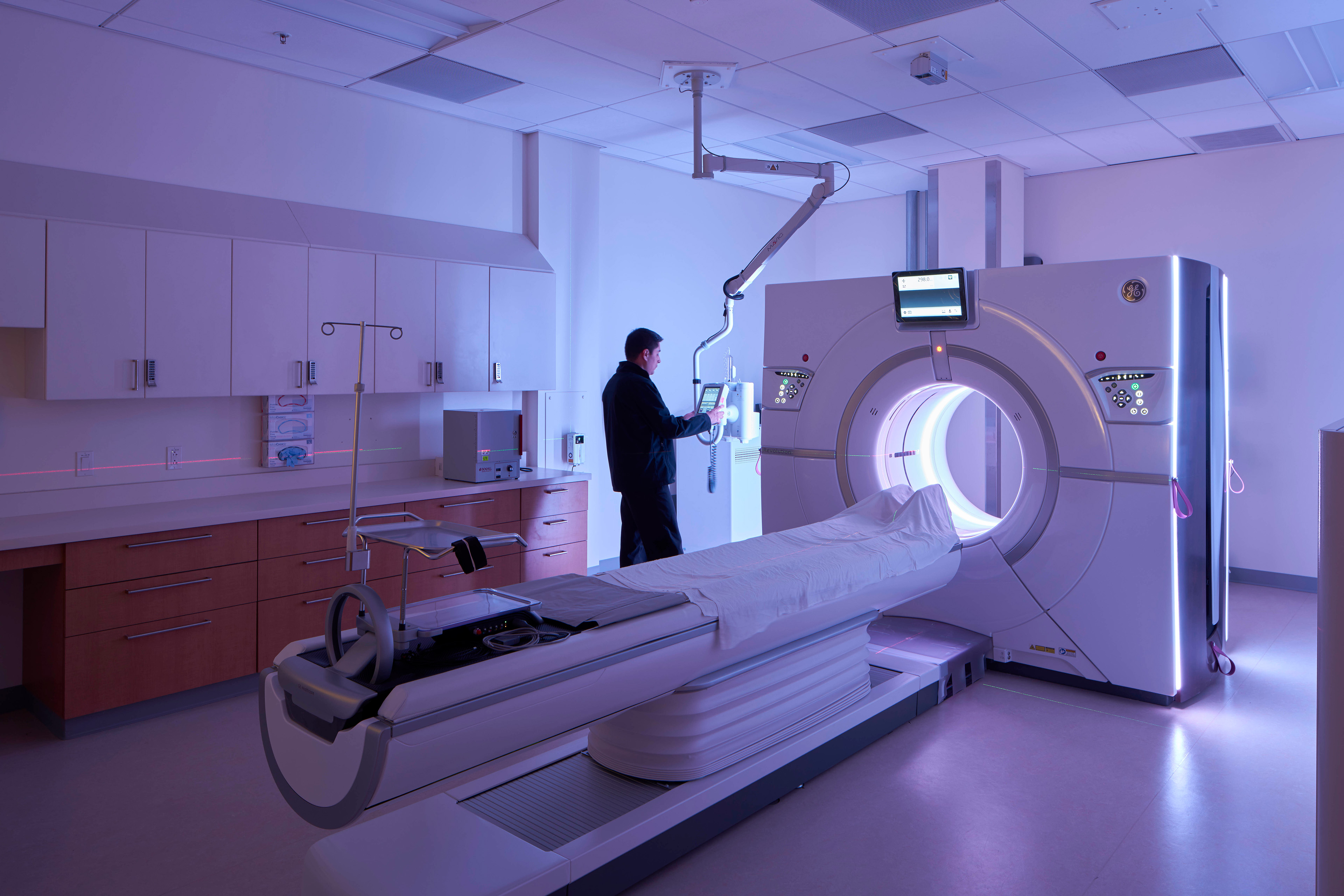 CT imaging suite illuminated with blue ambient lighting, featuring a scanner and patient table while a technician operates a ceiling-mounted control unit.