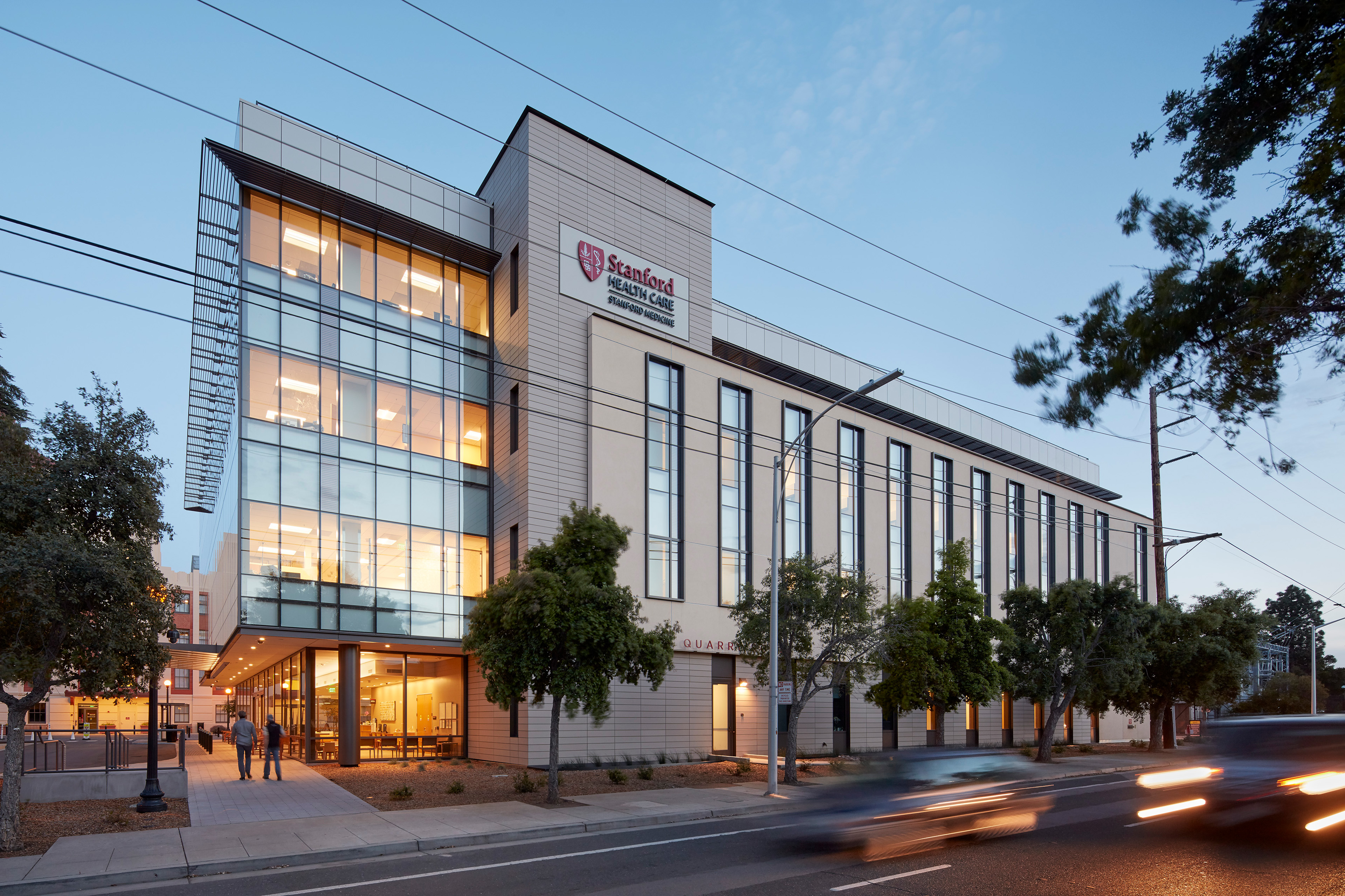 Dusk exterior of the Stanford Health Care Neuroscience Health Center with illuminated windows, prominent signage, street trees, and vehicles passing in front.