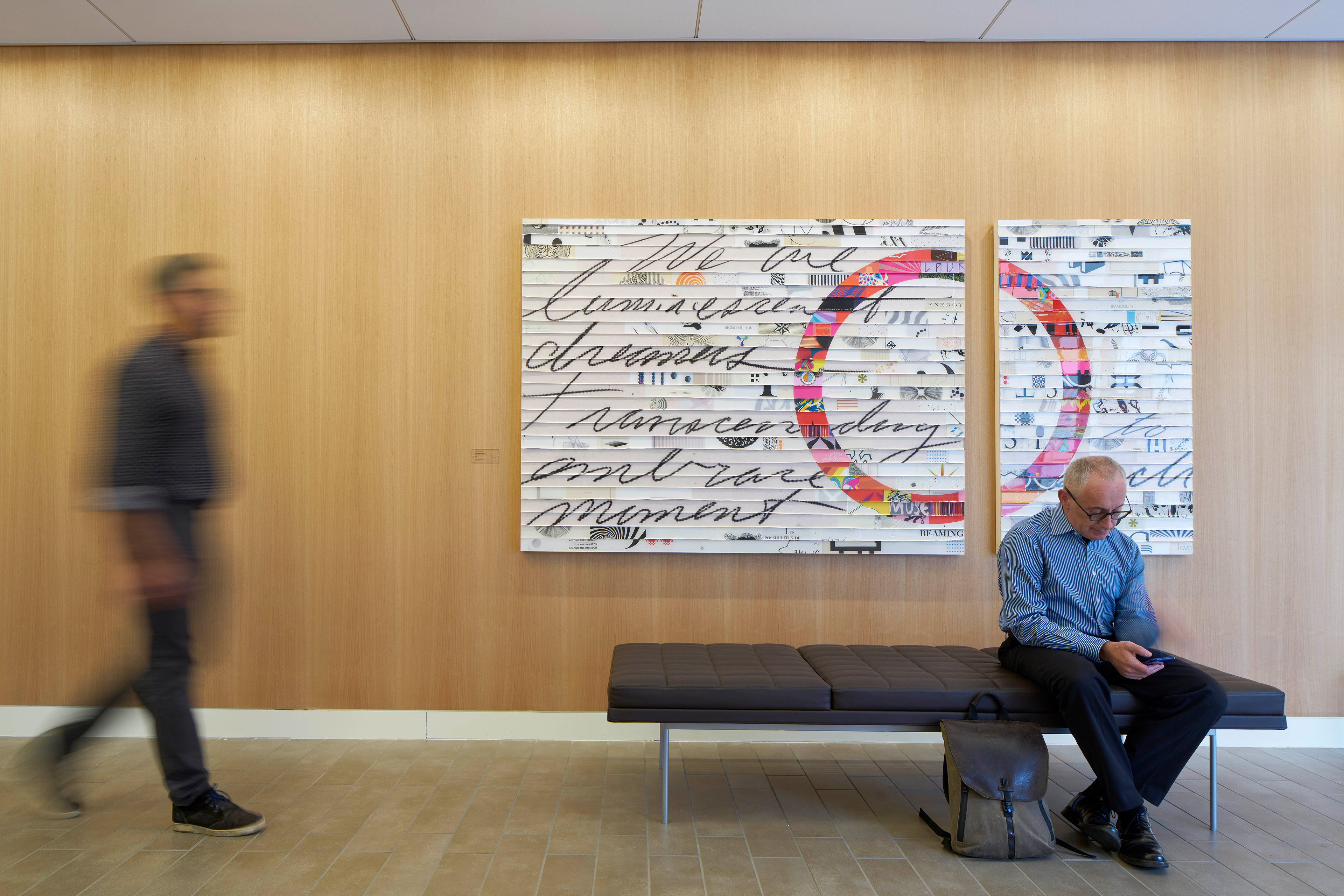 Wood-paneled lobby wall with large mixed-media artwork, a visitor seated on a bench using his phone, and another person walking past.