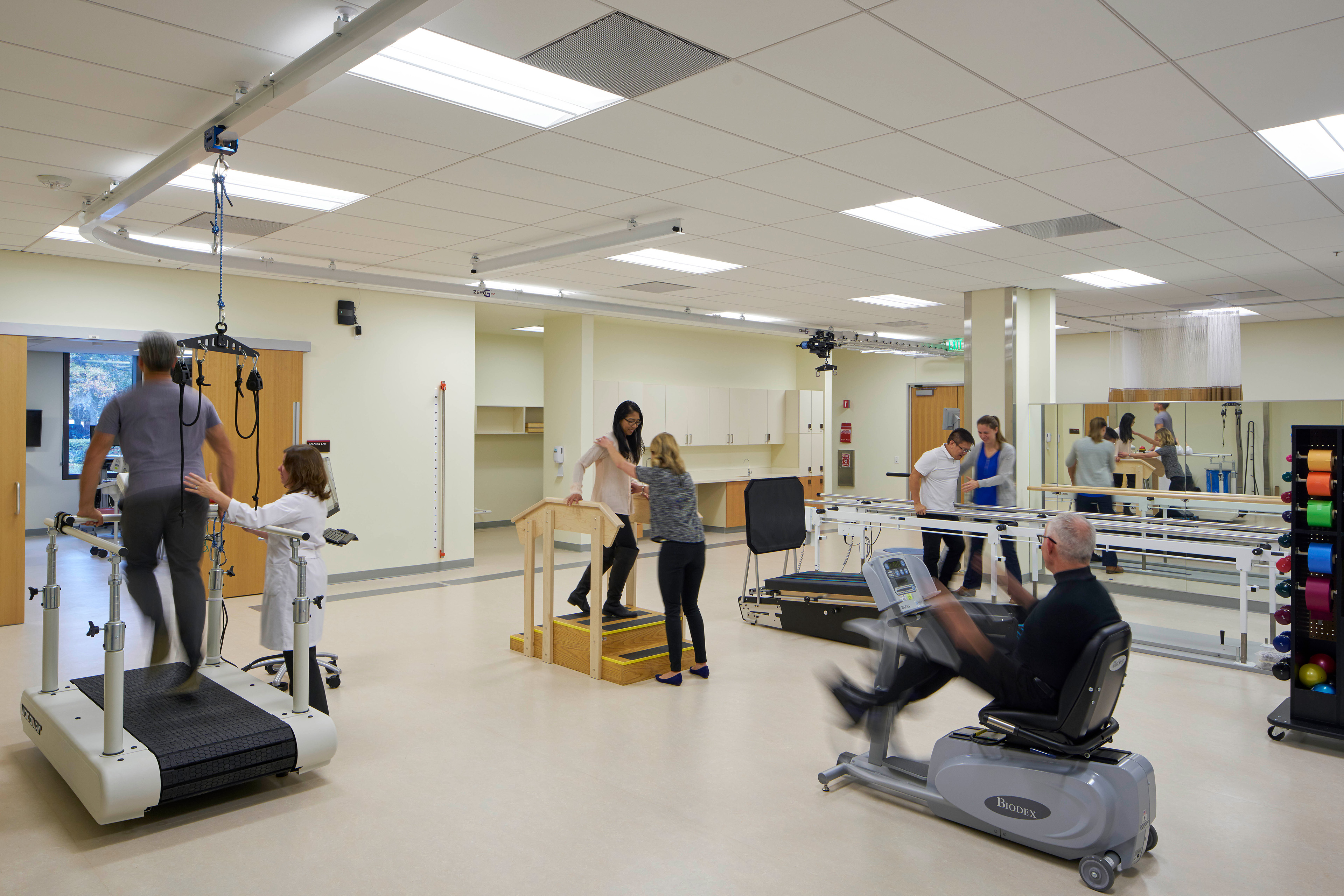 Rehabilitation gym with patients and clinicians using body-weight support equipment, parallel bars, training stairs, a recumbent exercise machine, and other therapy stations.