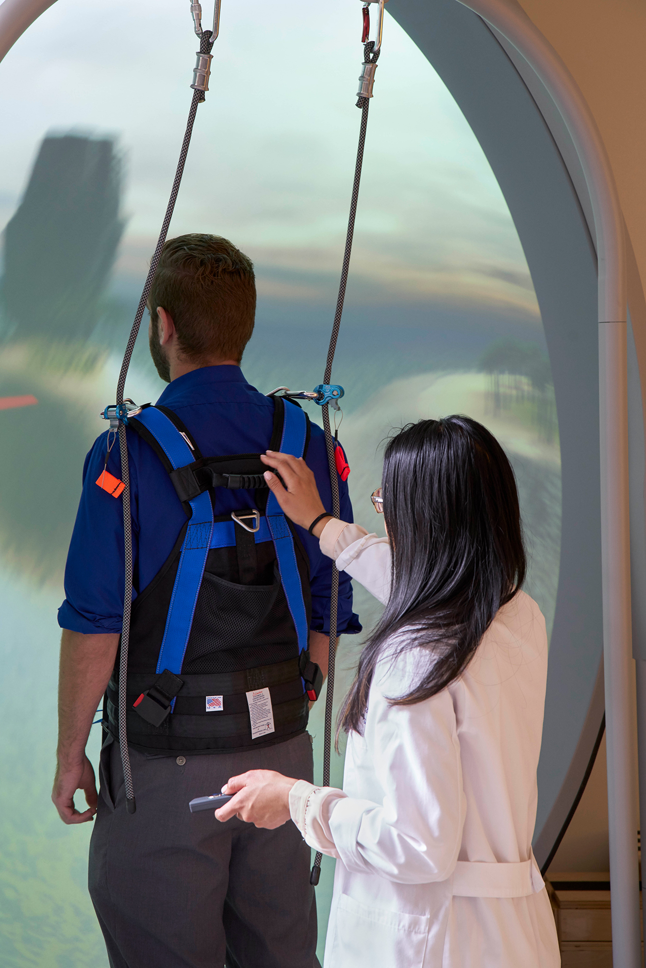 Clinician adjusting a safety harness on a patient standing in an immersive rehabilitation system with overhead support cables and a projected virtual environment.