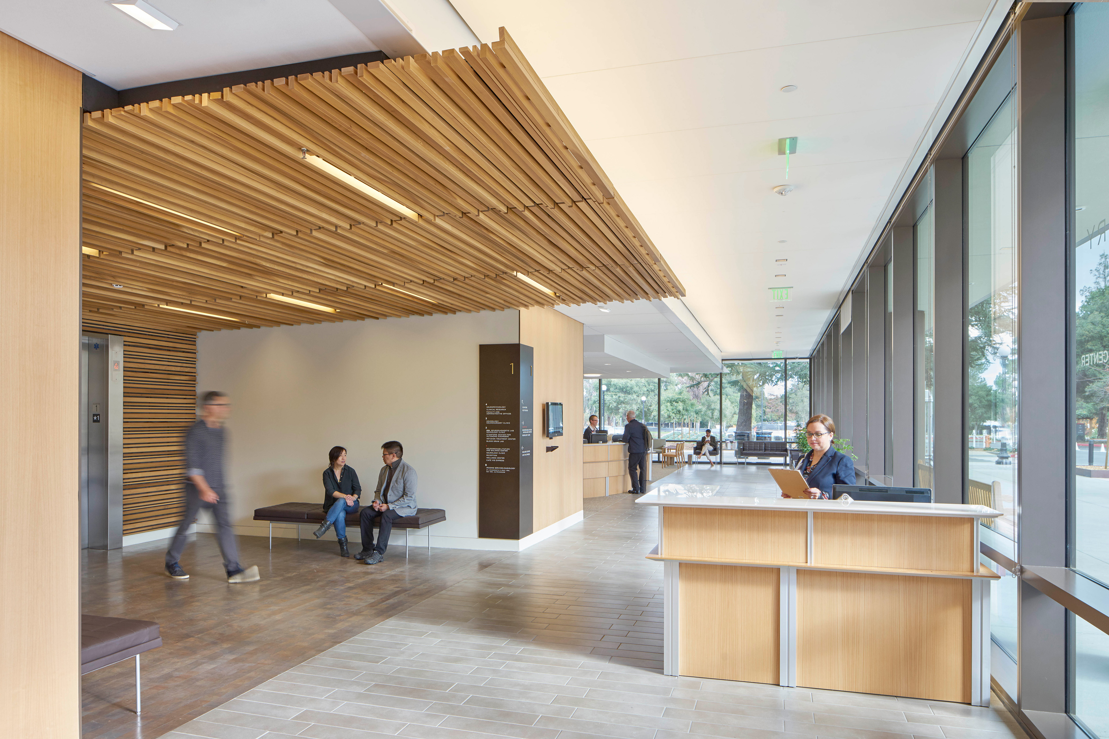 Bright healthcare lobby with a wood-slat ceiling, reception desk, built-in seating, floor-to-ceiling windows, and visitors and staff using the space.