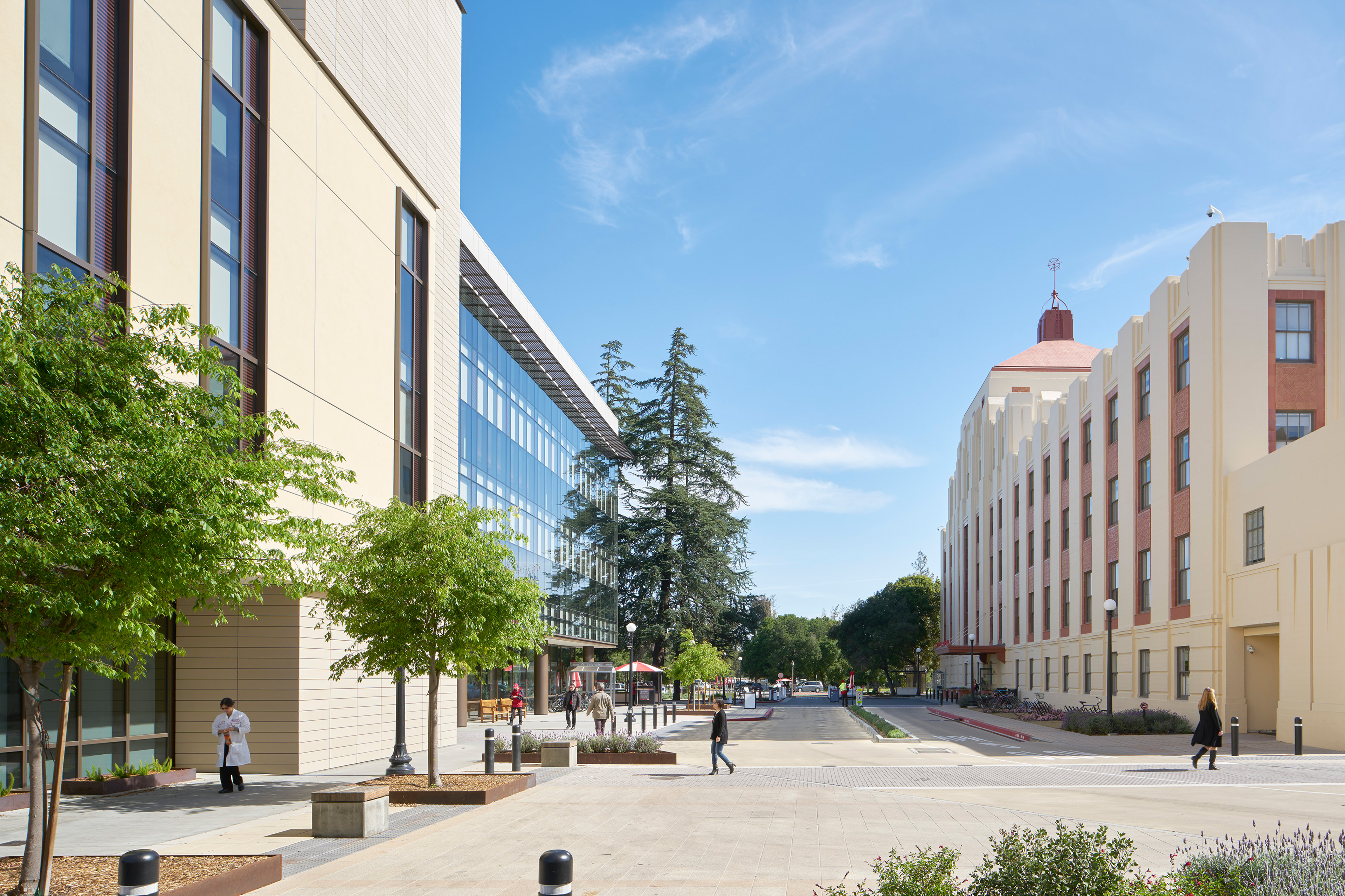 Pedestrian-oriented campus street between the modern Neuroscience Health Center and a historic hospital building, with trees, landscaping, and people walking through the plaza.
