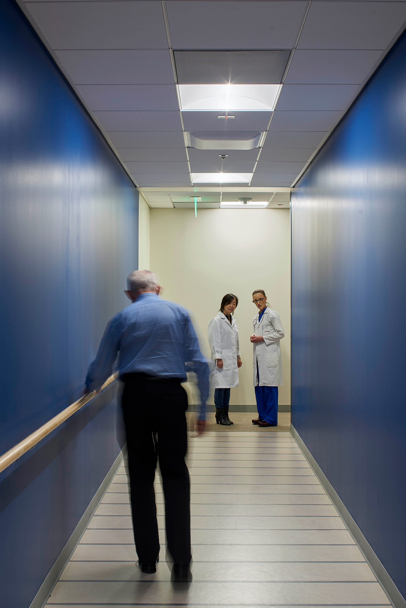 Blue-walled rehabilitation corridor with handrails, where a patient walks toward two clinicians in white coats at the far end.