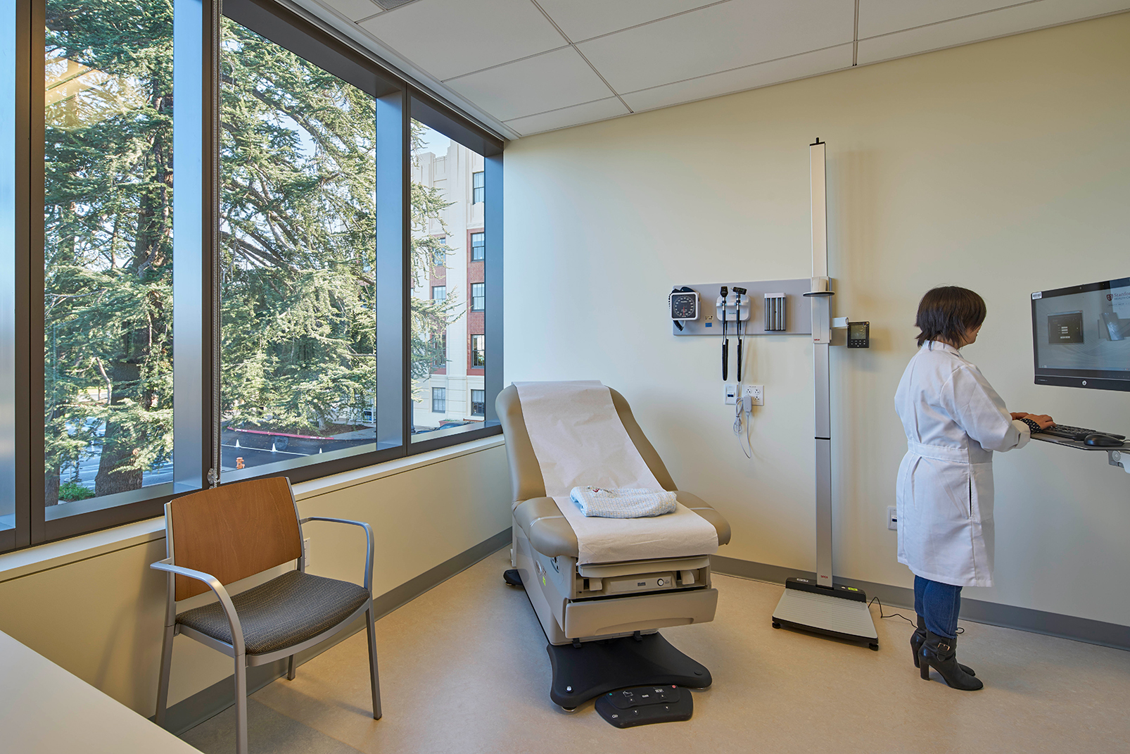 Clinical exam room with a large window, reclining exam chair, diagnostic equipment, accessible scale, guest seating, and a clinician entering information at a wall-mounted computer.