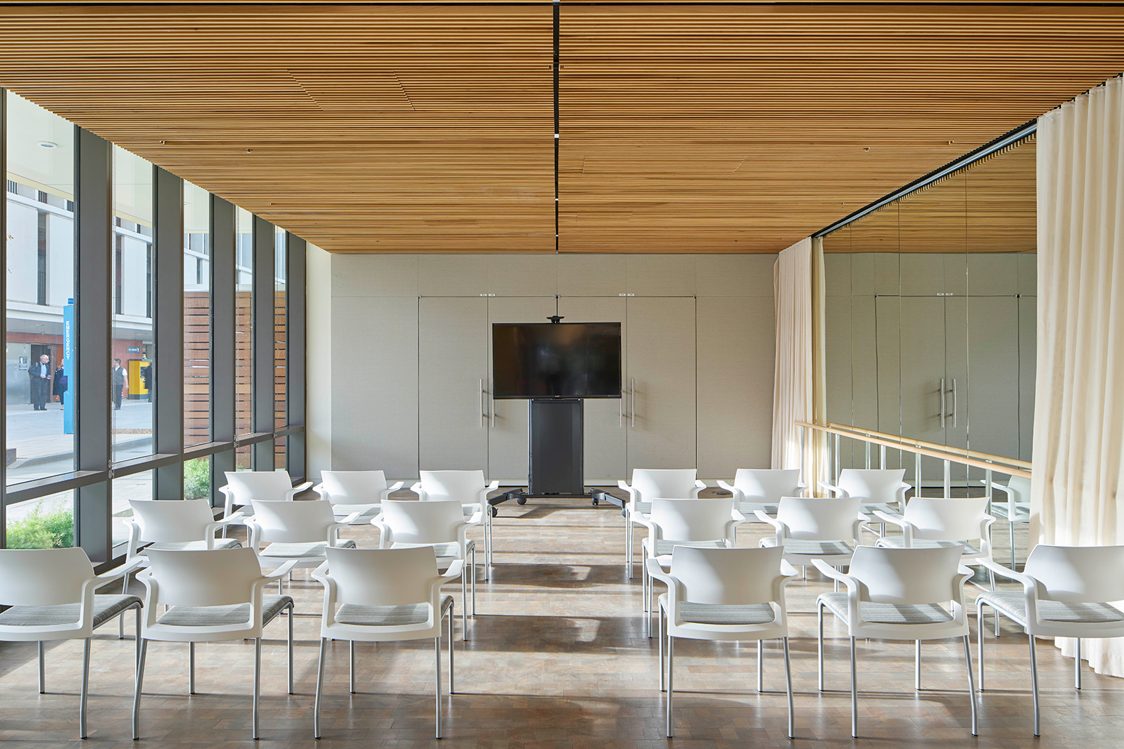 Flexible meeting and presentation room with rows of white chairs, a wood-slat ceiling, wall-mounted display, mirrored wall, and floor-to-ceiling windows.
