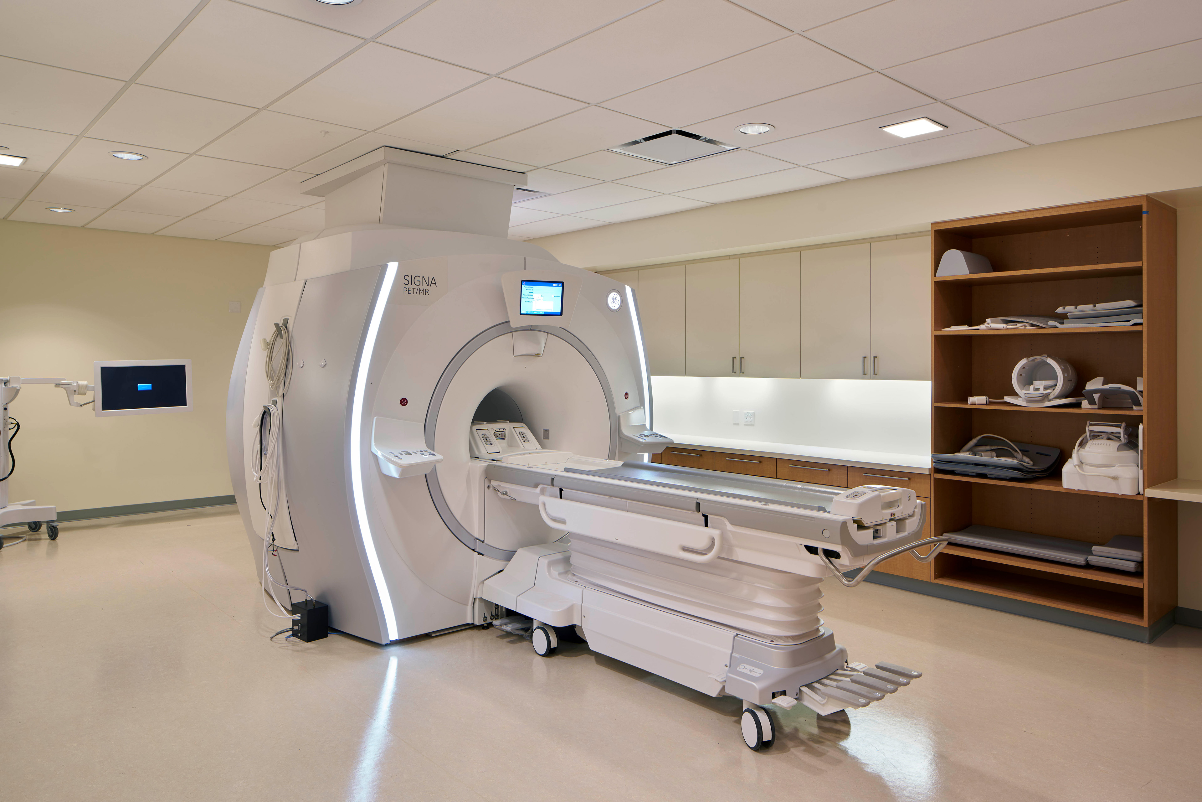 PET/MR imaging room with a GE Signa scanner, patient table, wall-mounted monitor, built-in cabinetry, and organized imaging accessories.