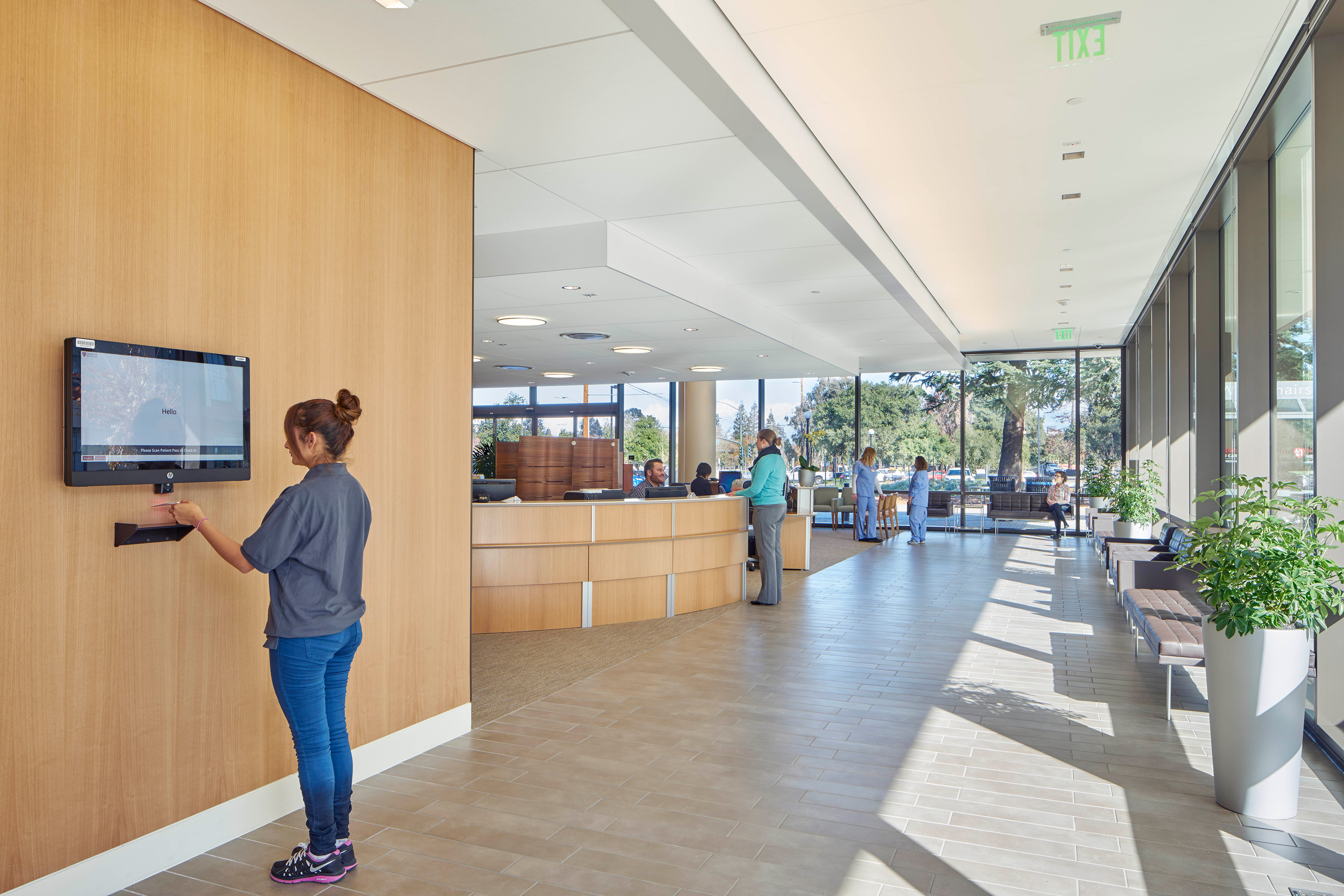 Sunlit healthcare lobby with a self-service check-in screen, curved reception desk, floor-to-ceiling windows, indoor plants, and staff and visitors using the space.