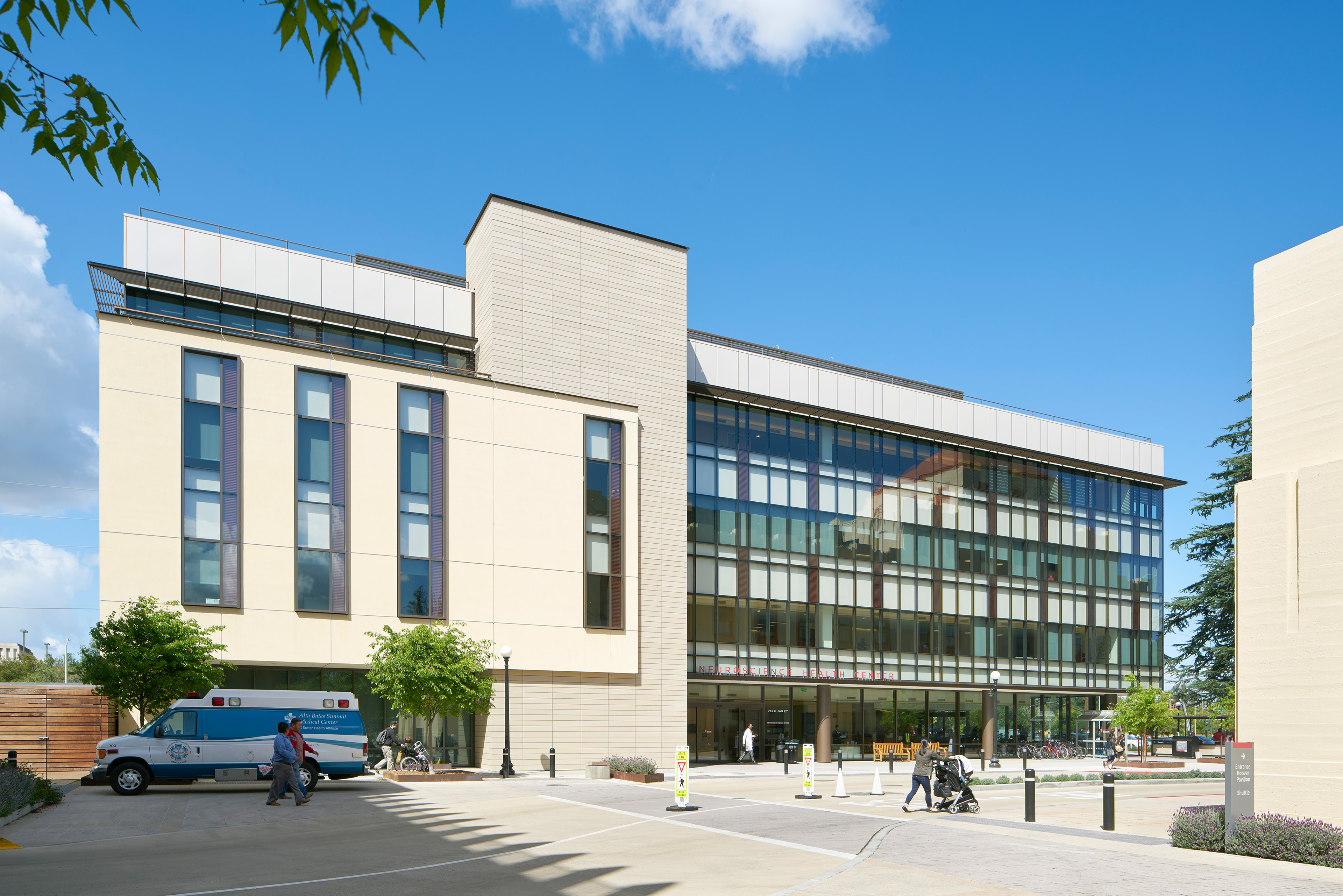 Daytime exterior of the Stanford Health Care Neuroscience Health Center, showing its glass-and-stone façade, main entrance, landscaped plaza, an ambulance, and pedestrians crossing in front.