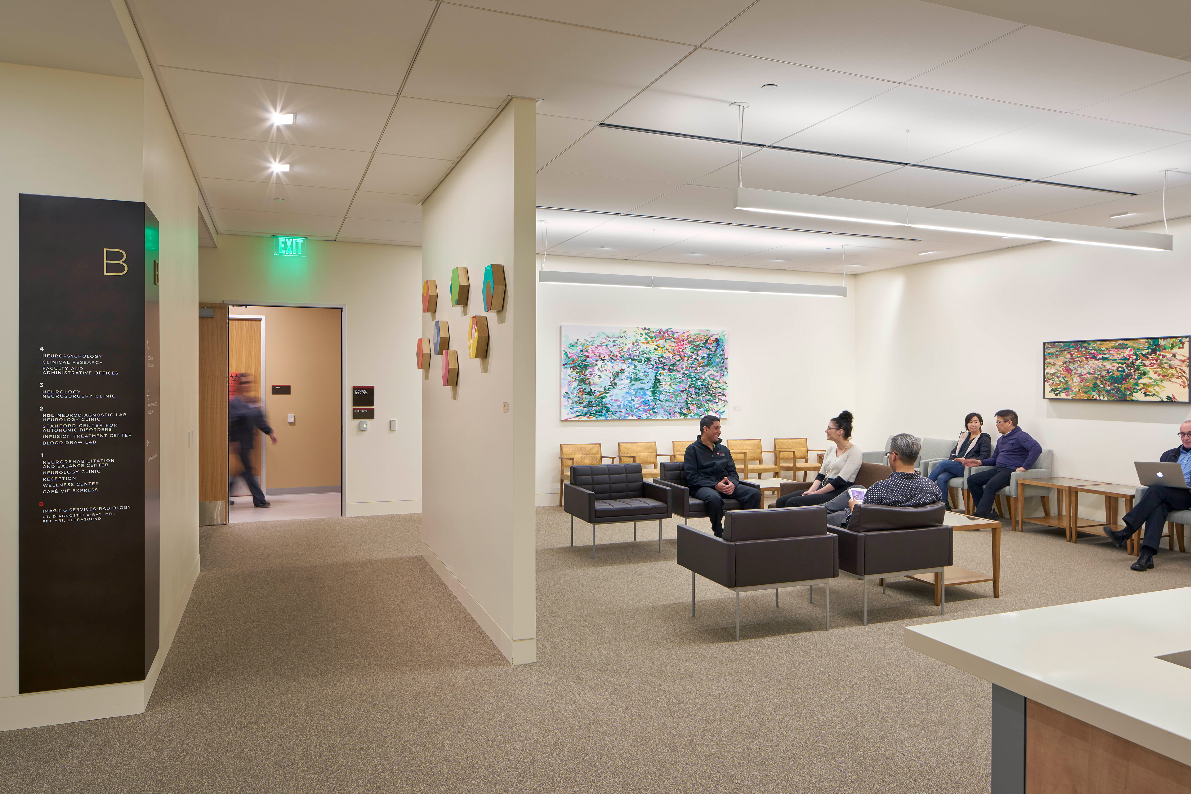 Healthcare waiting area with lounge seating, colorful artwork, a floor directory, and patients and visitors seated beneath suspended linear lights.