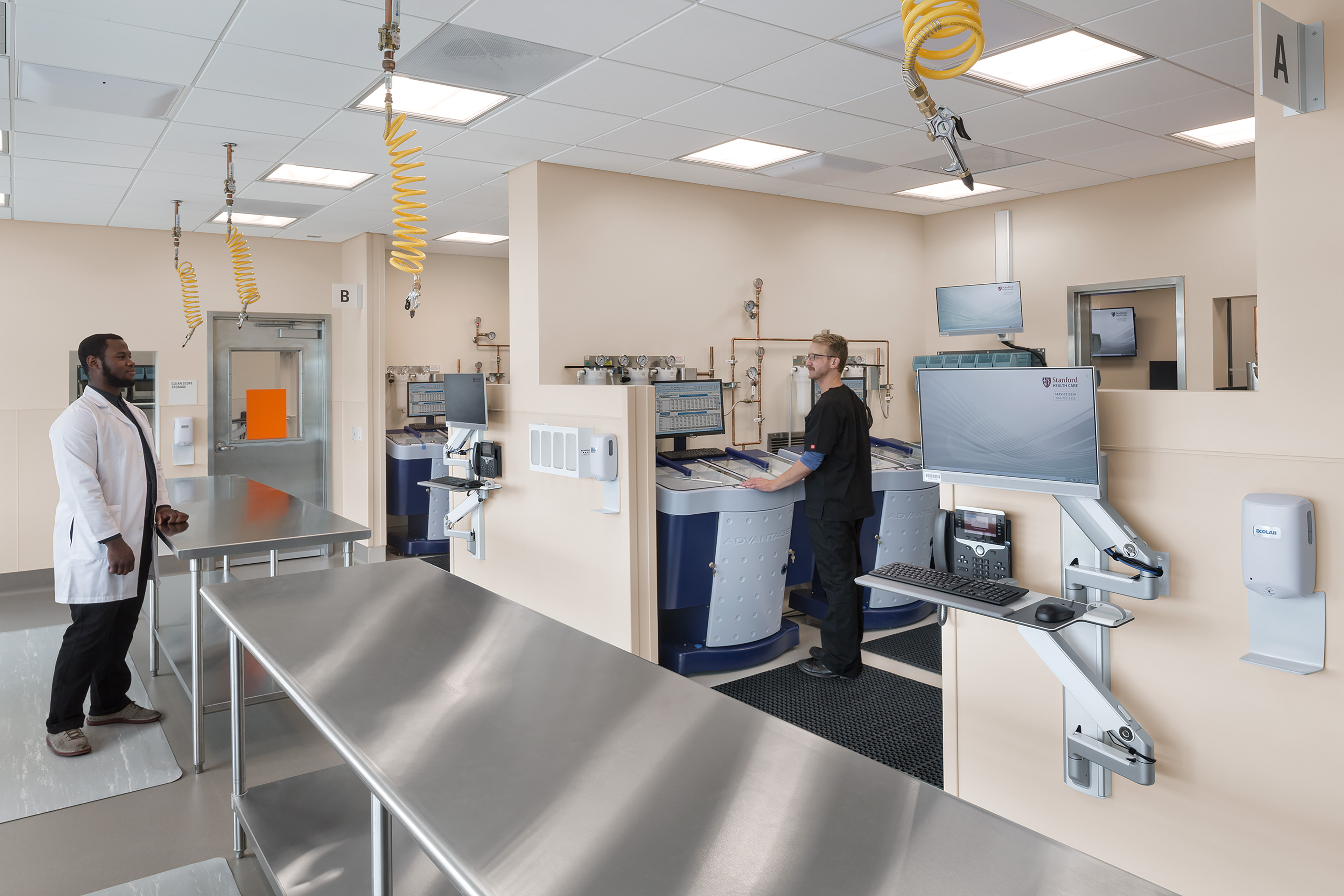 Medical equipment processing room with stainless steel worktables, automated scope reprocessors, wall-mounted computer stations, and two staff members.