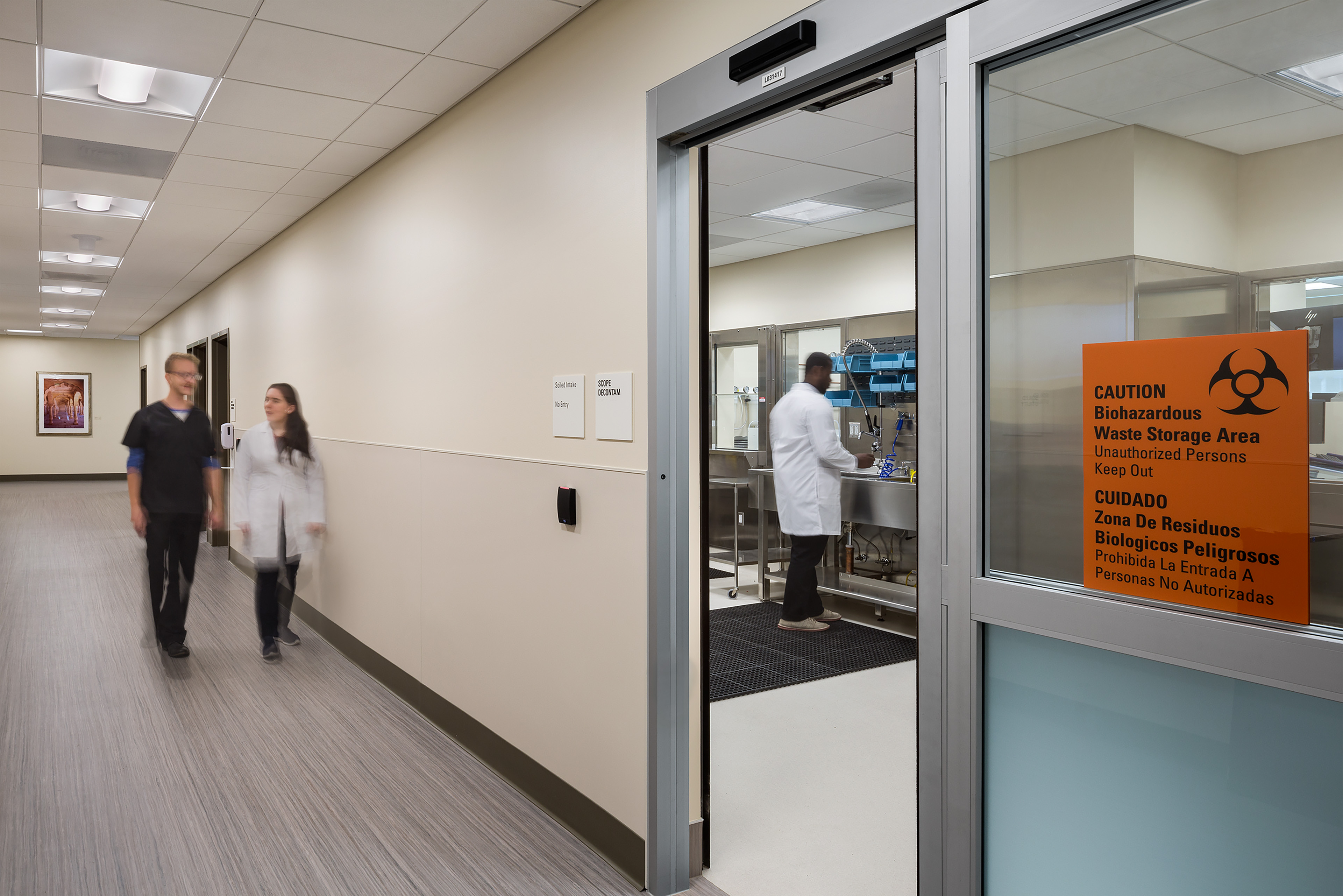 Clinical support corridor beside a biohazardous waste storage and scope decontamination room, with staff working and moving through the space.