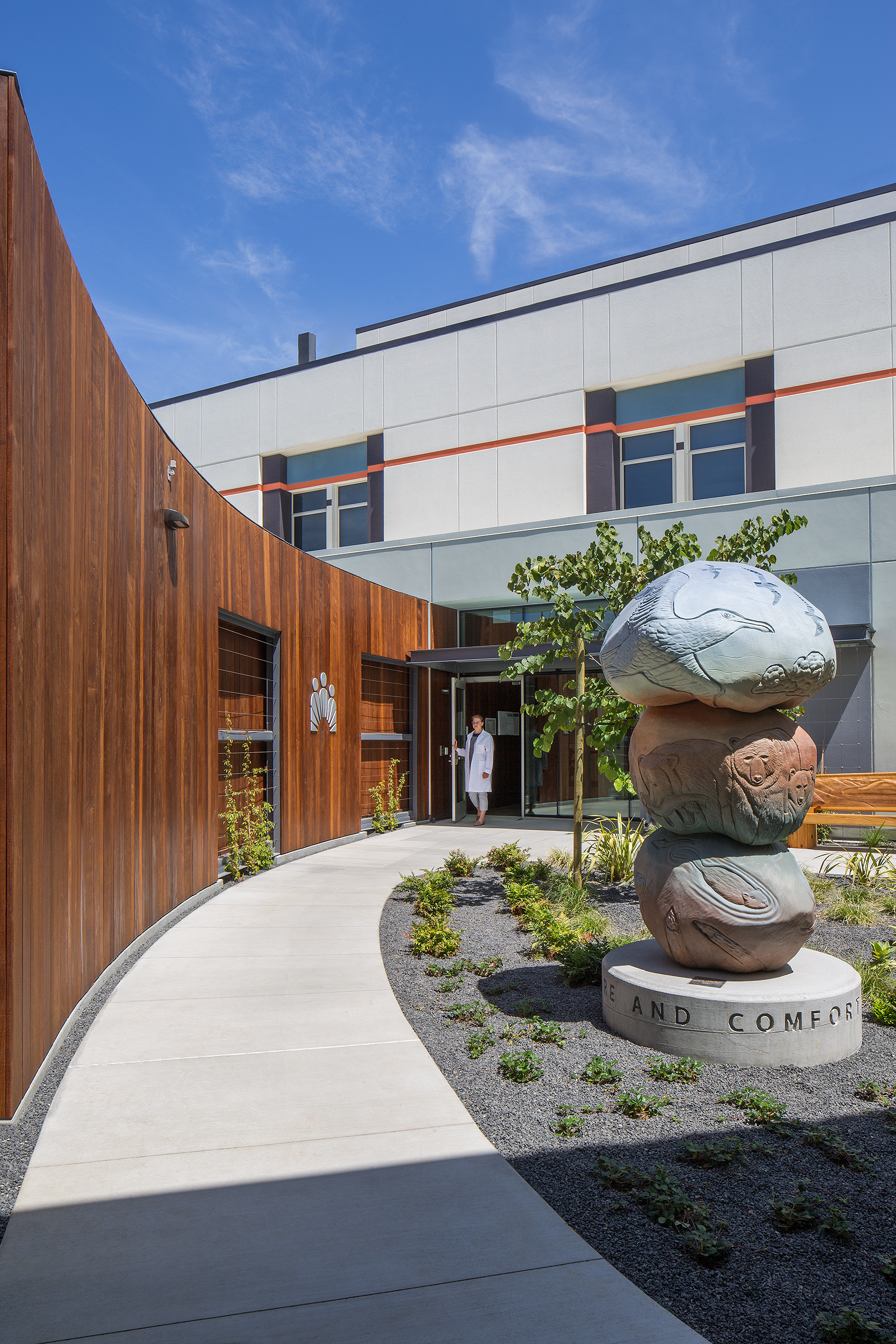 Landscaped hospital courtyard with curved wood walls, a sculptural stone artwork, and a clinician standing near the entrance.