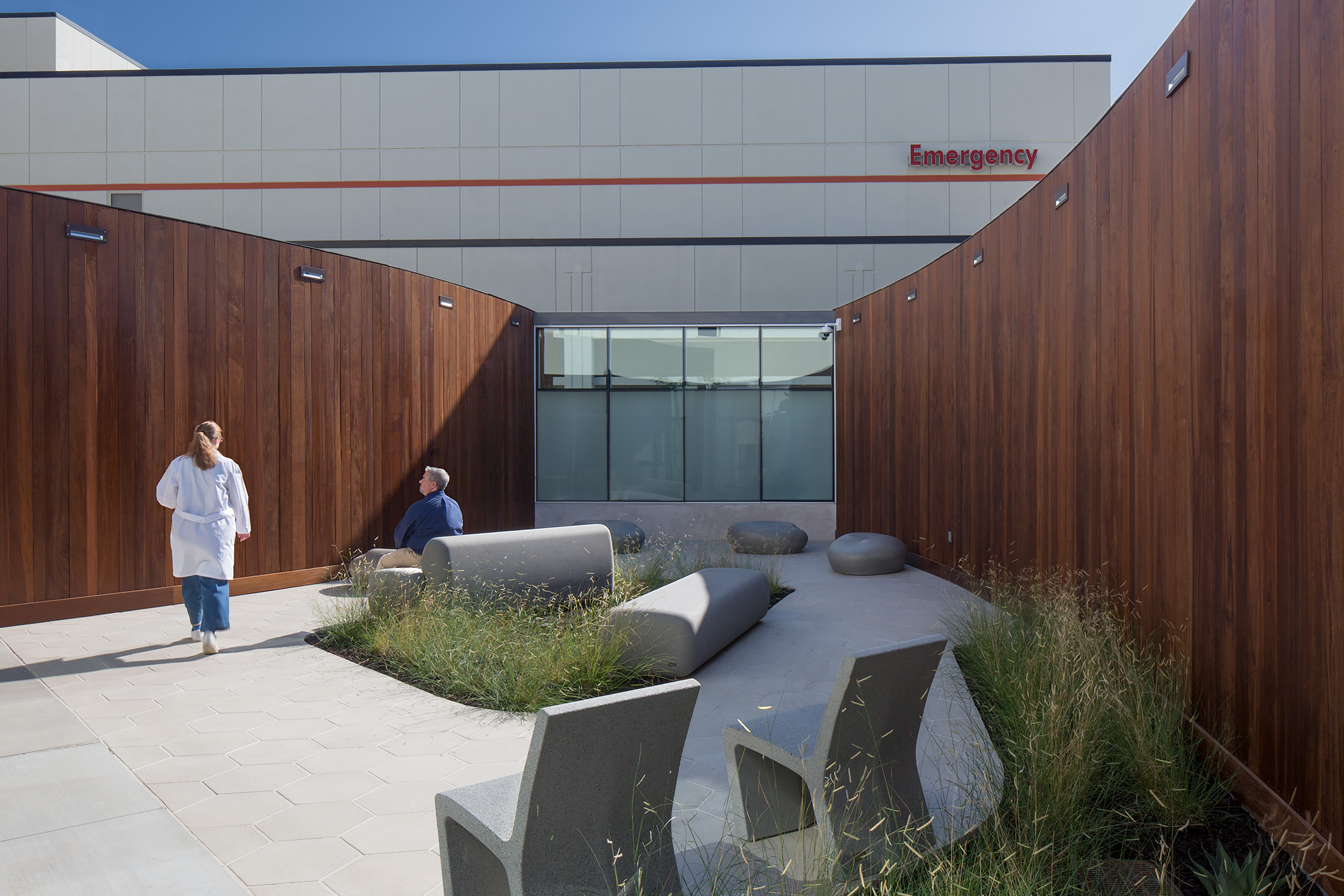Outdoor healing courtyard beside an emergency department, with wood-clad walls, landscaped grasses, sculptural seating, and a clinician and visitor using the space.