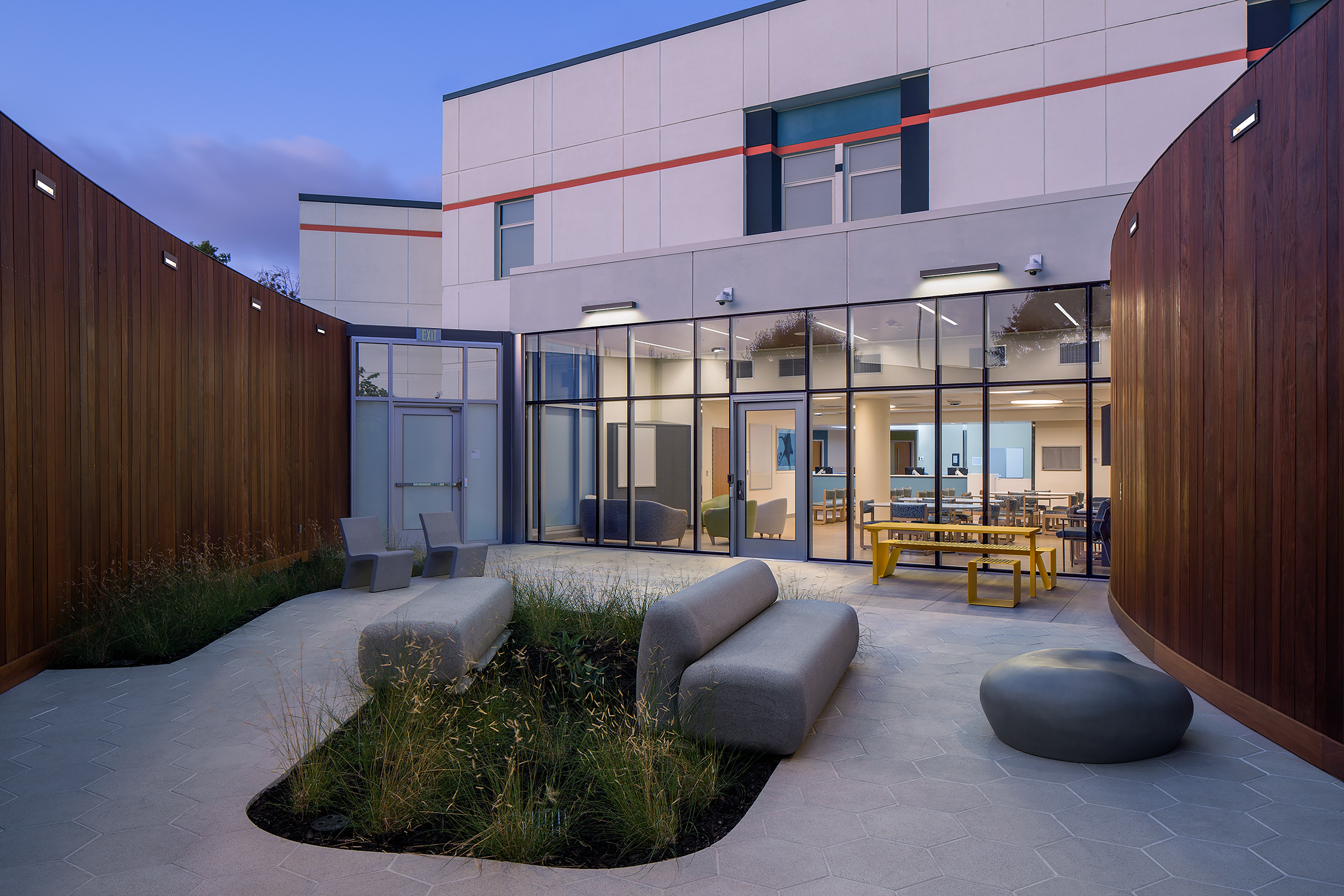 Landscaped hospital courtyard at dusk with curved wood walls, sculptural seating, and glass doors opening to a brightly lit lounge.