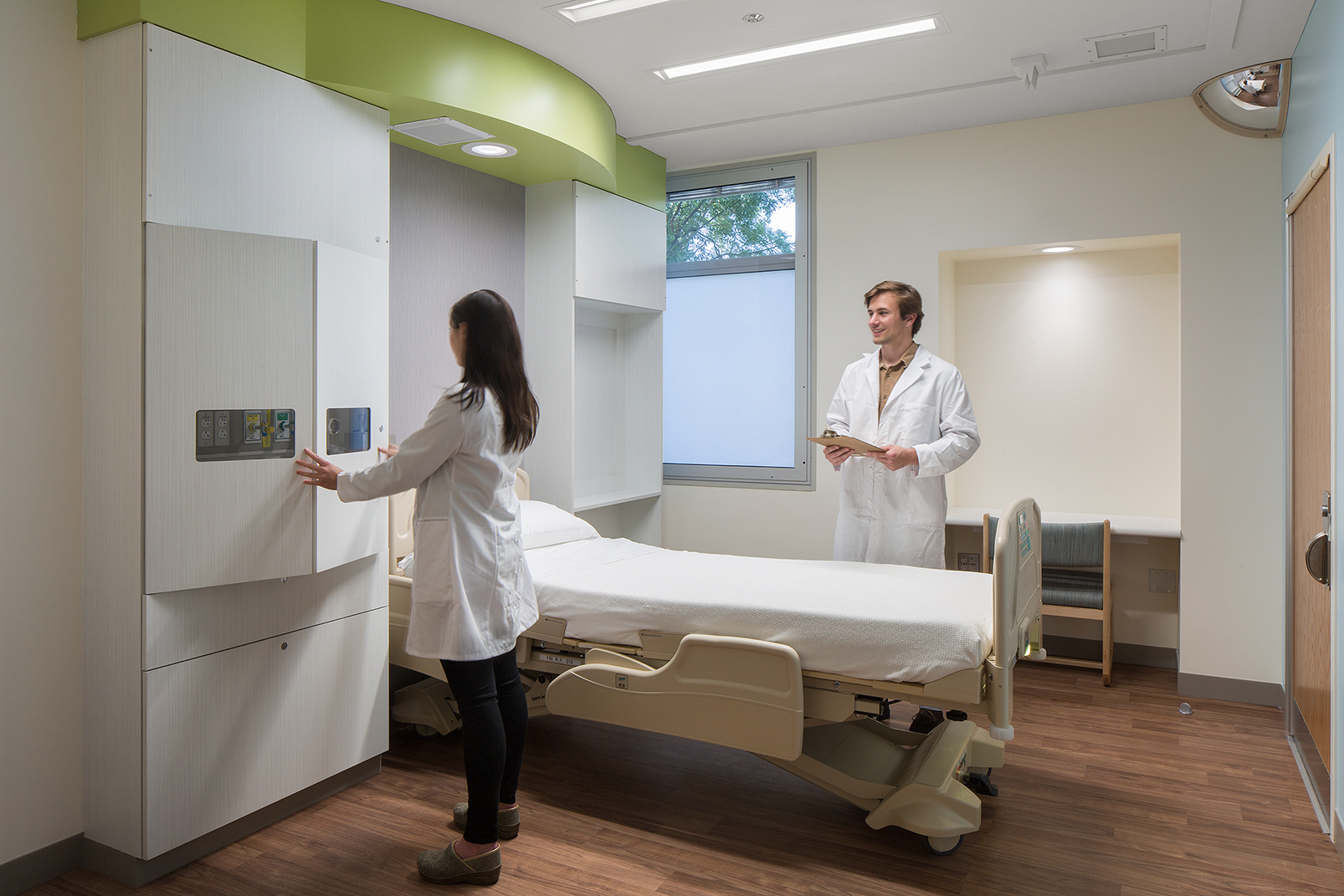 Hospital patient room with a bed, built-in storage, and two clinicians reviewing the room’s equipment and layout.