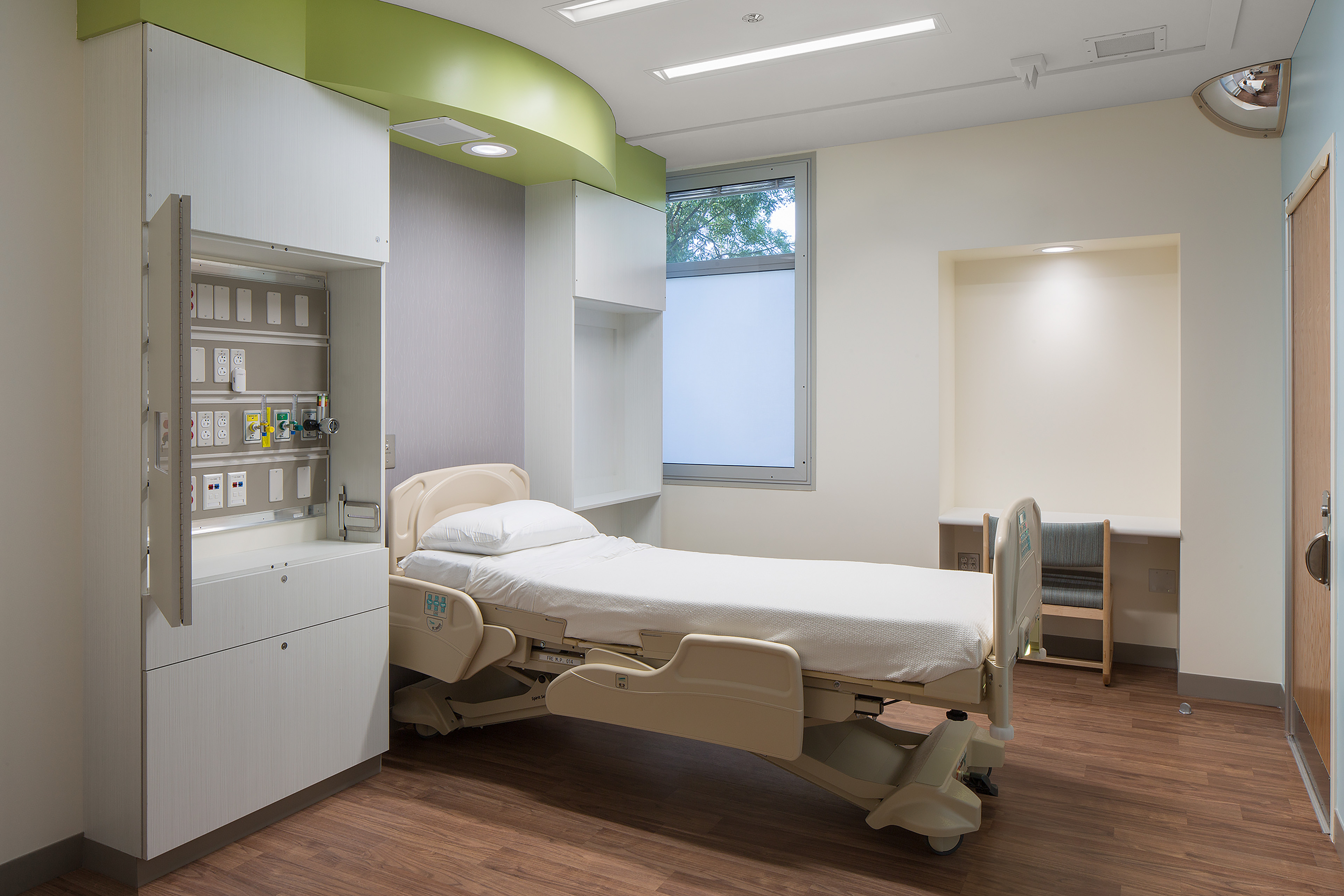 Behavioral health patient room with a hospital bed, concealed medical services, durable built-in cabinetry, wood-look flooring, a partially obscured window, and a recessed desk area.