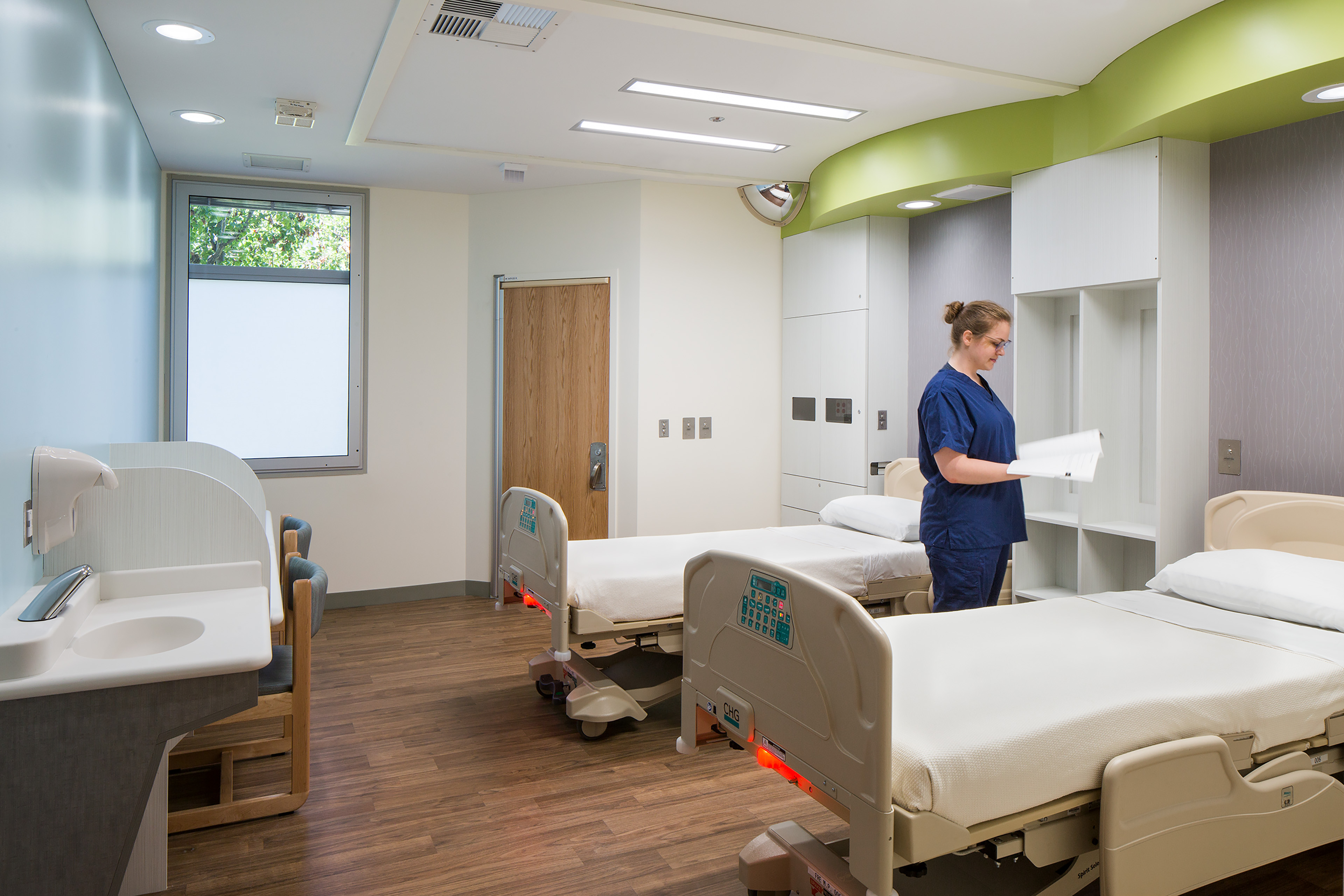 Shared hospital room with three patient beds, built-in storage, a sink, and a clinician reviewing paperwork.