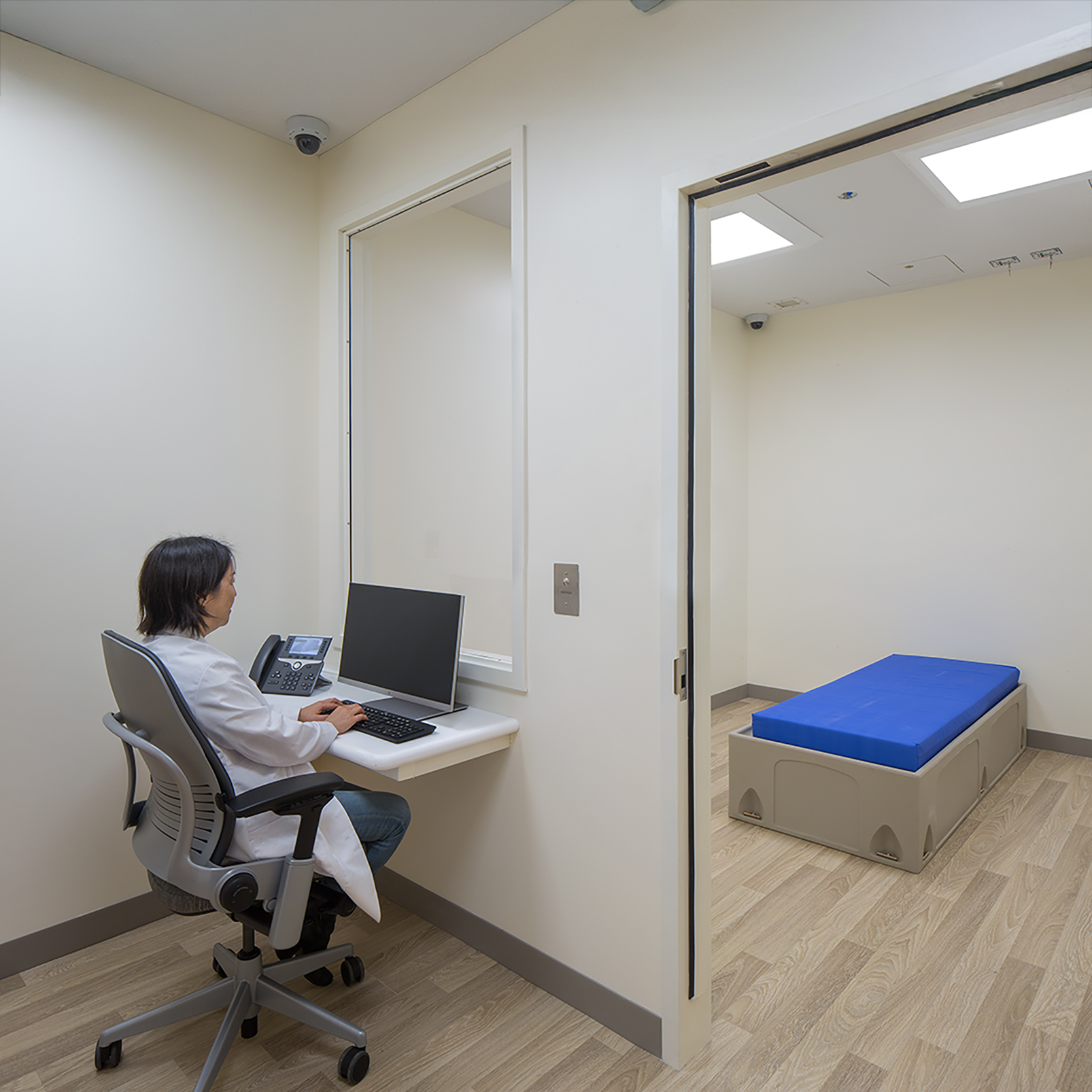 Clinical observation room with a staff workstation, interior viewing window, security cameras, and a padded platform in the adjacent patient space.