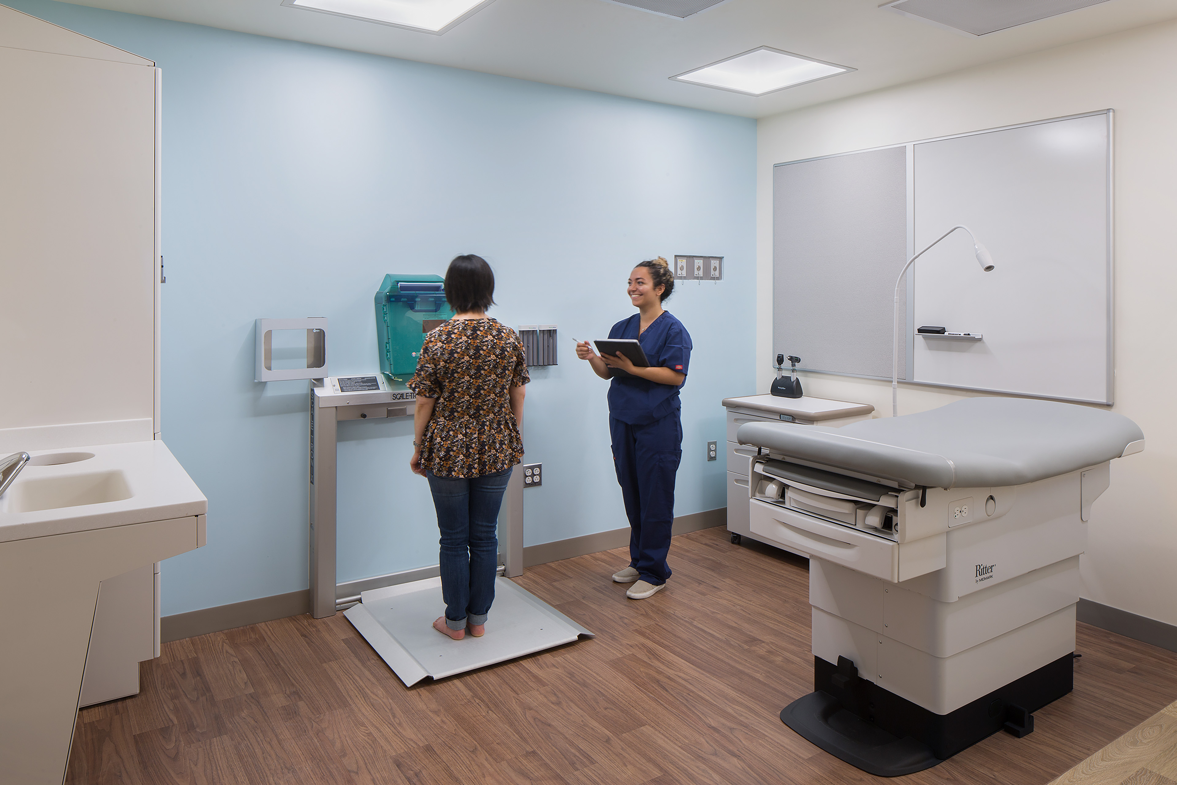 Clinical exam room with a patient standing on a medical scale while a clinician records information on a tablet beside an exam table.