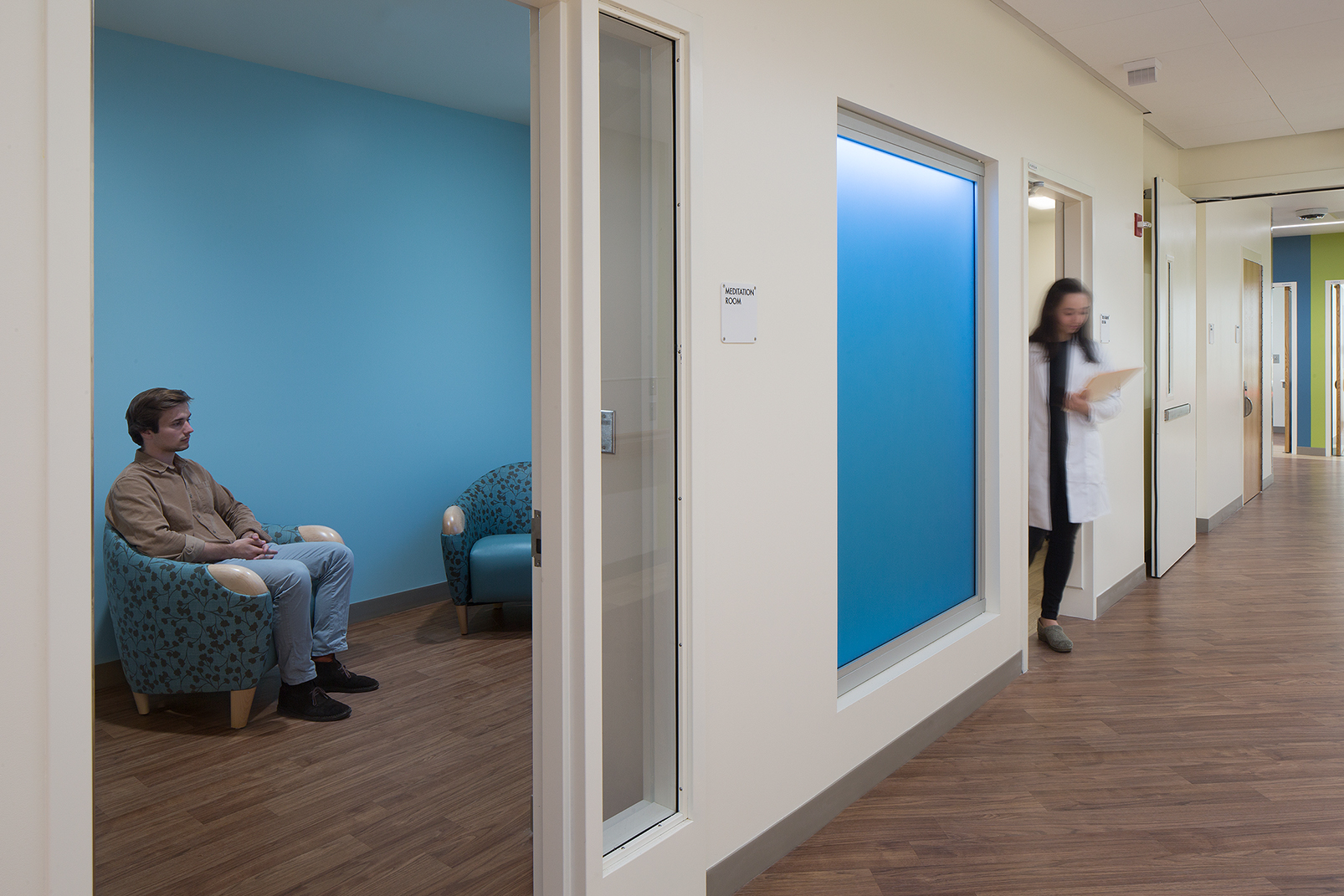 Medication room with blue accent walls and upholstered seating, where a patient sits quietly while a clinician walks through the adjacent healthcare corridor.