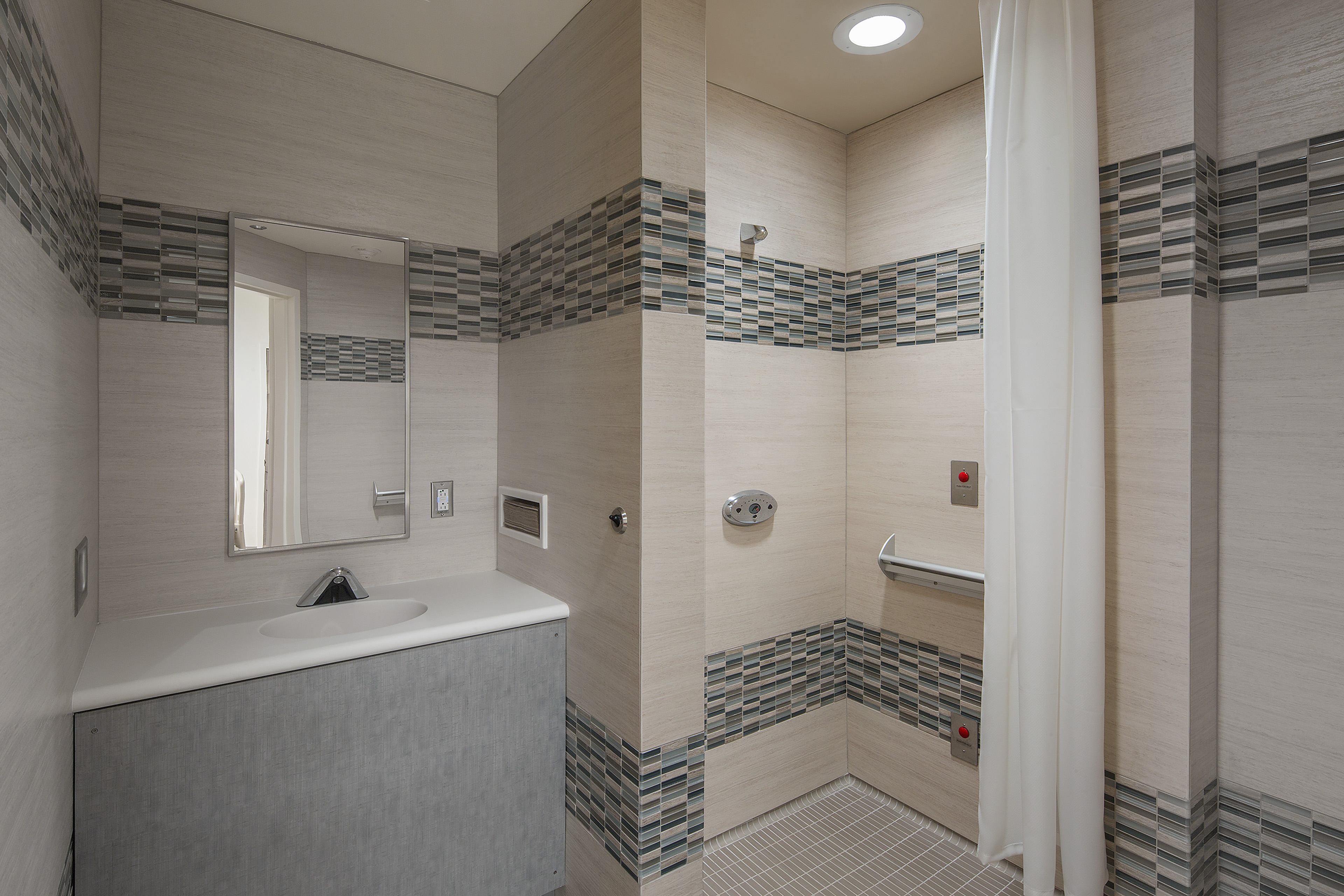 Accessible patient bathroom with a sink, mirror, roll-in shower, grab bars, privacy curtain, and blue-gray mosaic tile accents.