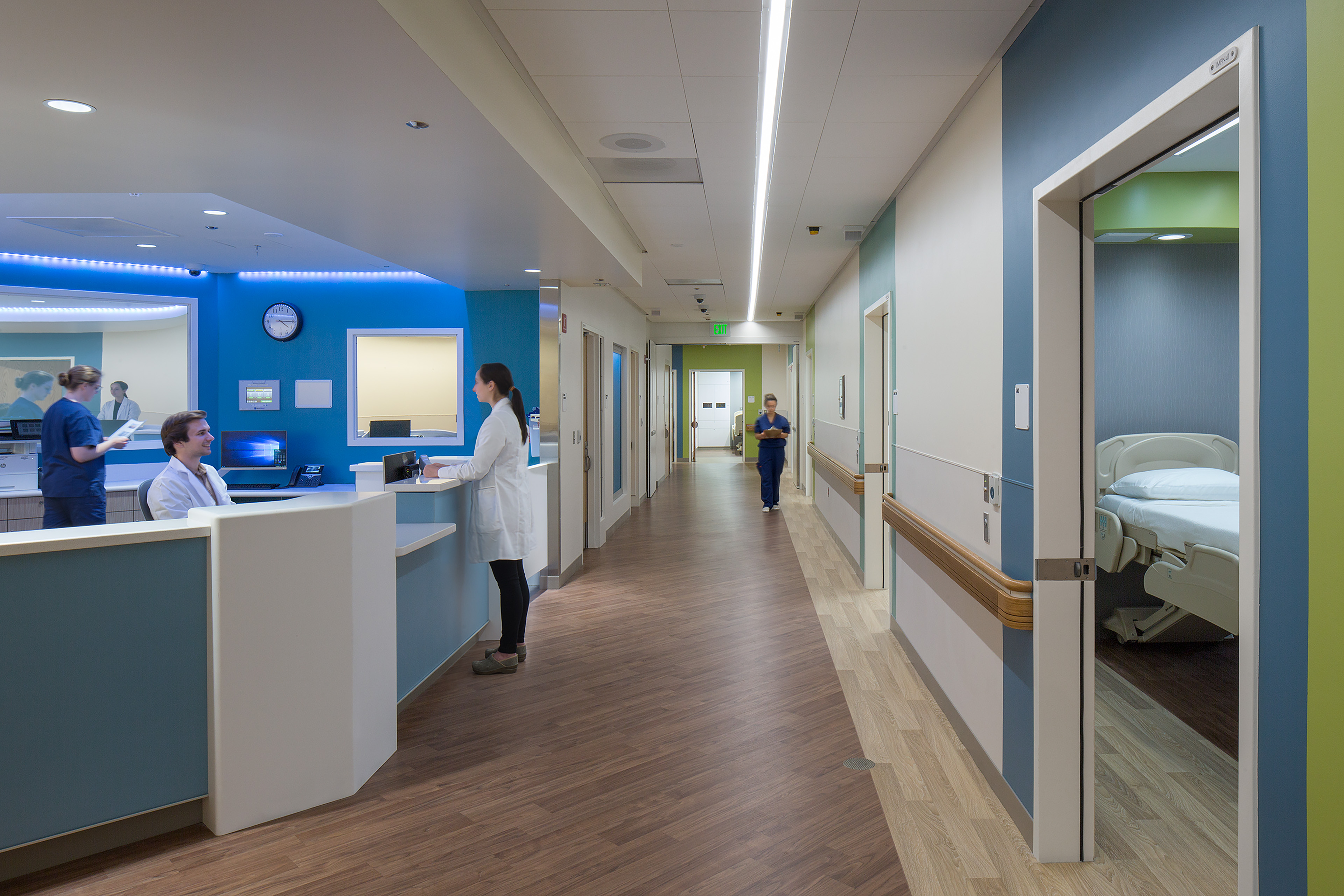 Hospital corridor with a central nurses’ station, patient rooms, and several clinicians working or walking through the brightly lit space.