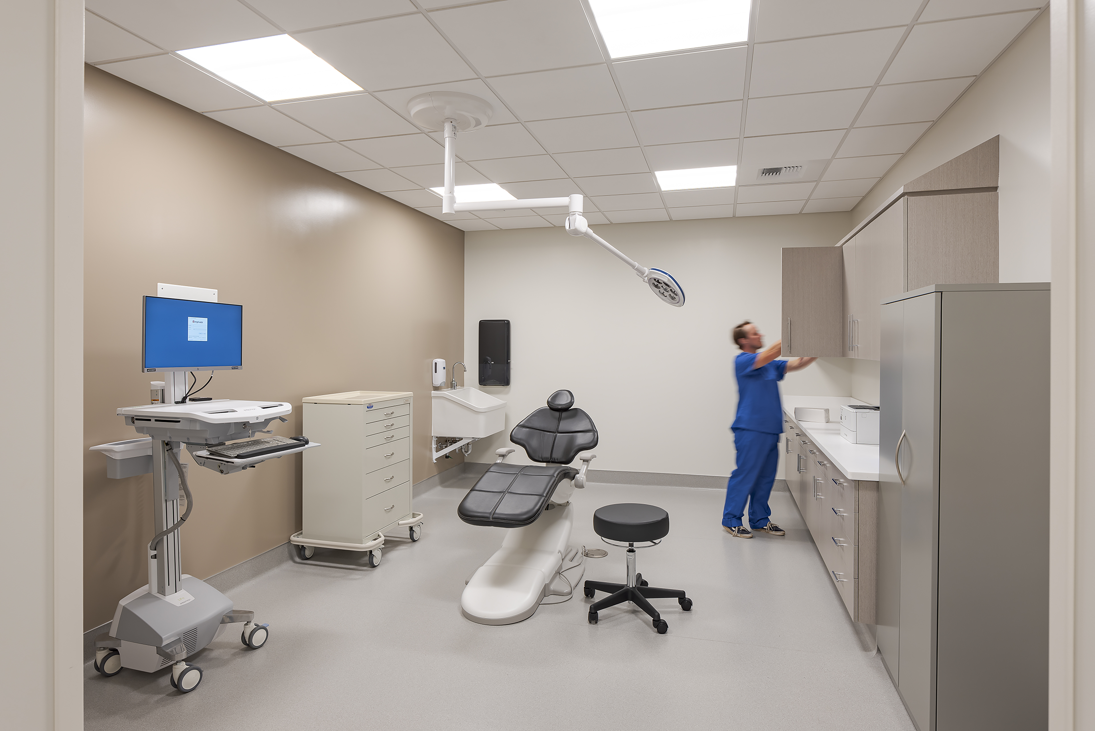 Ophthalmic procedure room with an exam chair, ceiling-mounted surgical light, mobile computer workstation, medical storage, sink, and a clinician preparing the space.