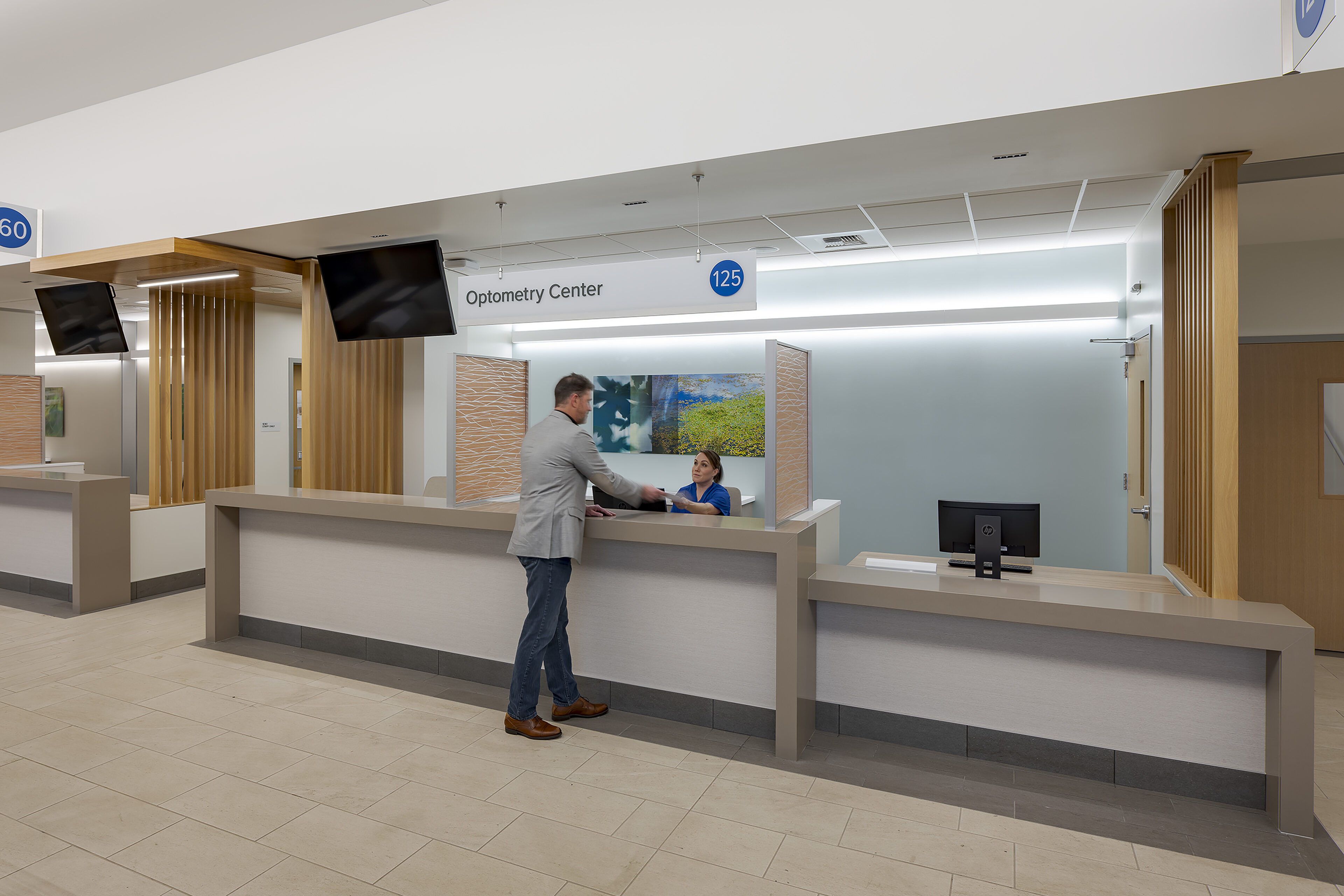 Optometry Center reception desk with warm wood accents, overhead signage, and a staff member assisting a visitor.