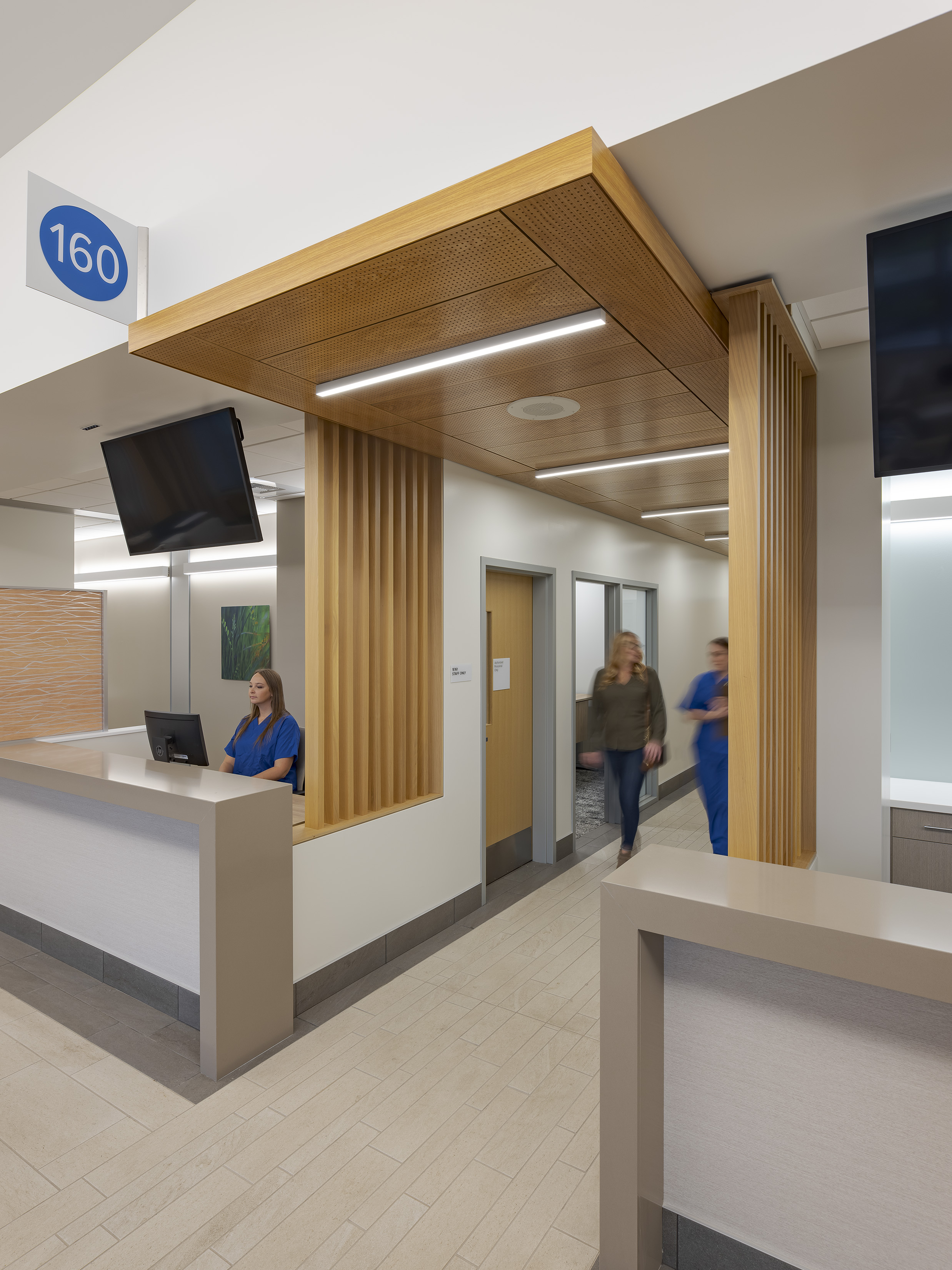 Reception and check-in area with numbered signage, warm wood canopy and slatted details, wall-mounted screens, and staff and visitors moving through the space.