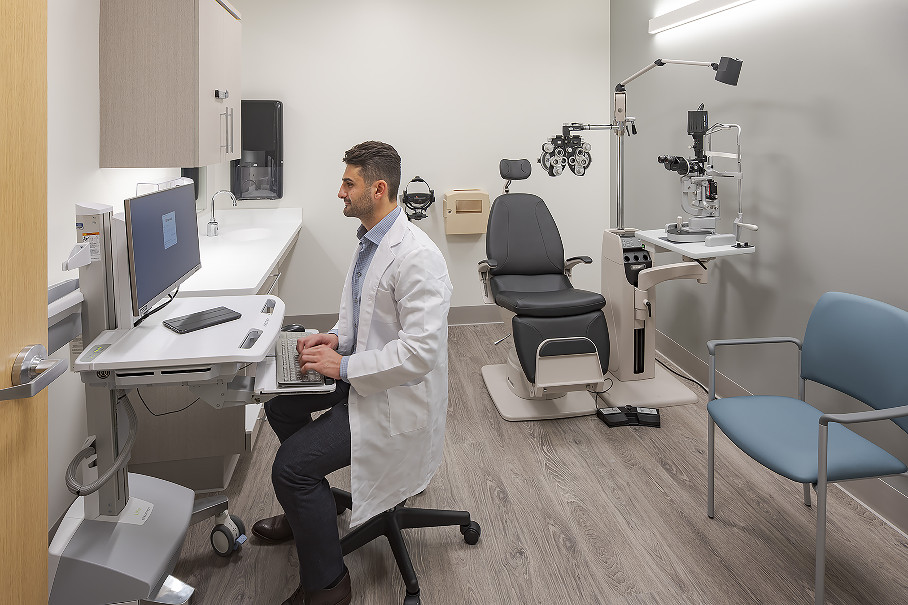 Eye exam room with ophthalmic equipment, an exam chair, accessible workstation and sink, and a physician entering information at a computer.